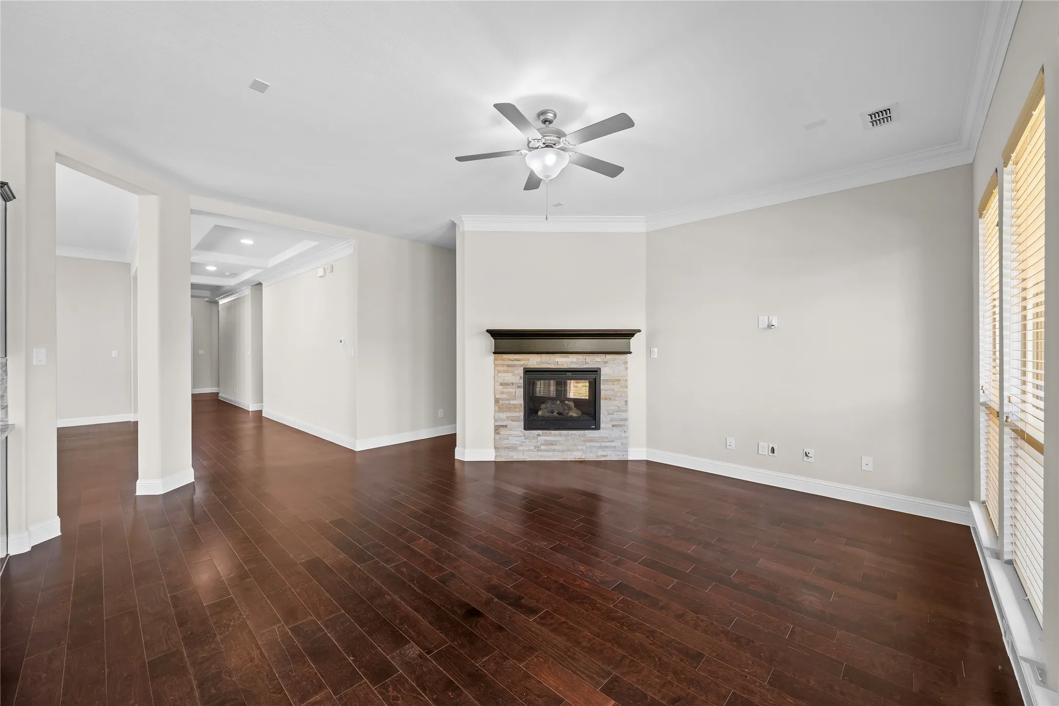 Wood-finish flooring, corner fireplace with stone surround, ceiling fan, crown molding, and tray ceiling in hallway
