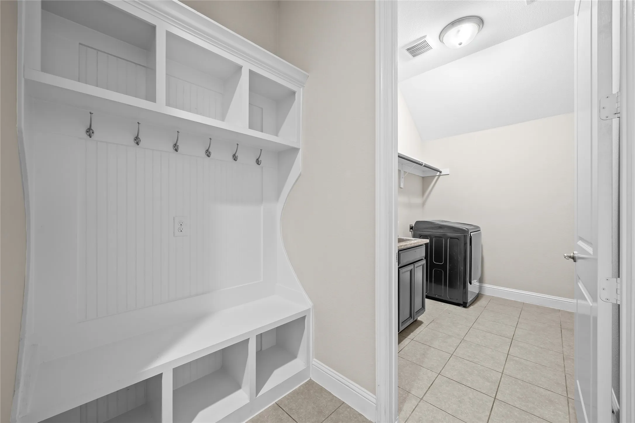 Dedicated mudroom and laundry space featuring built-in cabinetry with open shelving, multiple wall-mounted hooks for storage, tile flooring, and direct access to the utility area with washer and dryer connections