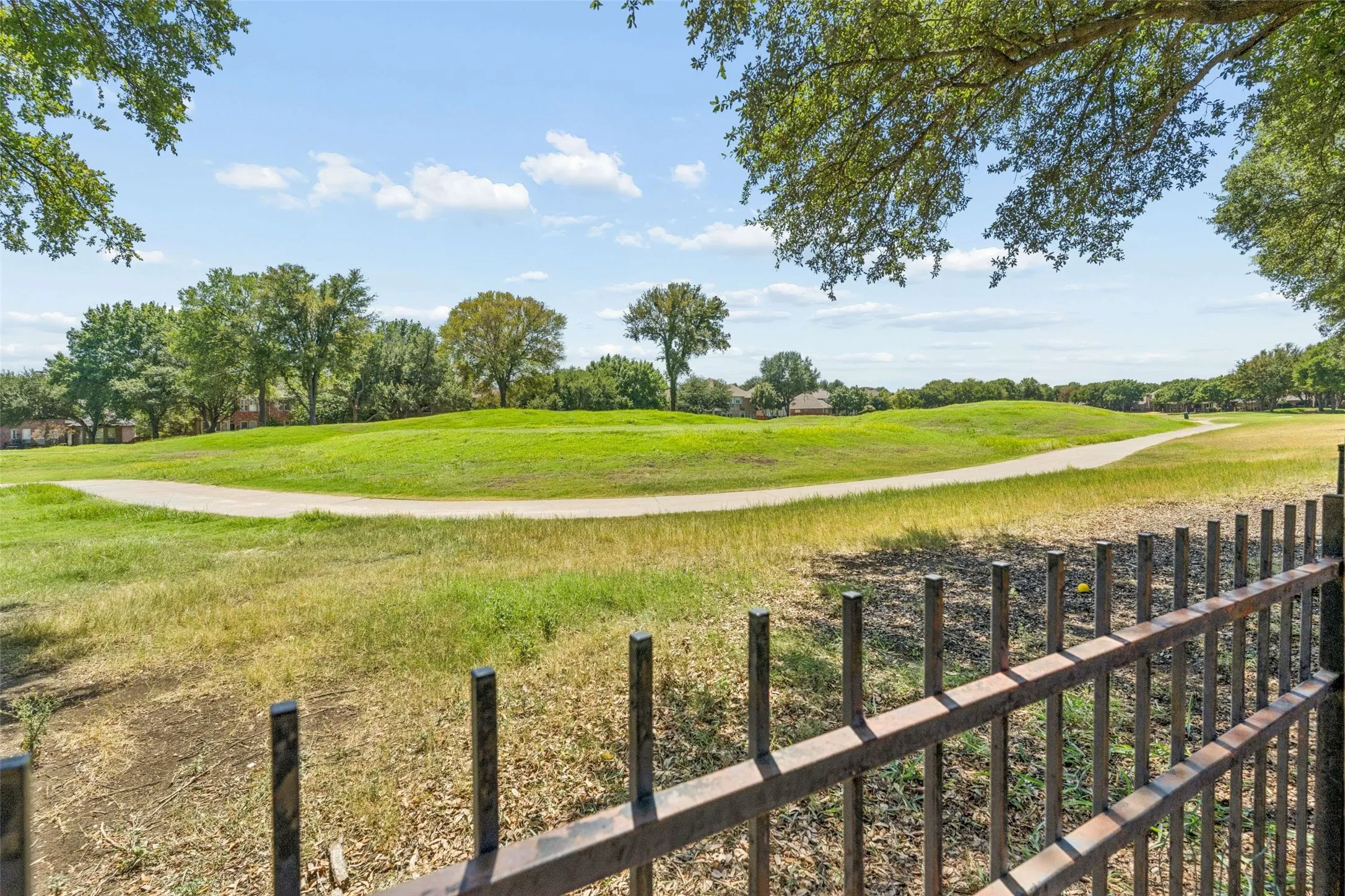 Expansive green space featuring rolling hills and a paved pathway, bordered by mature trees and a black metal fence