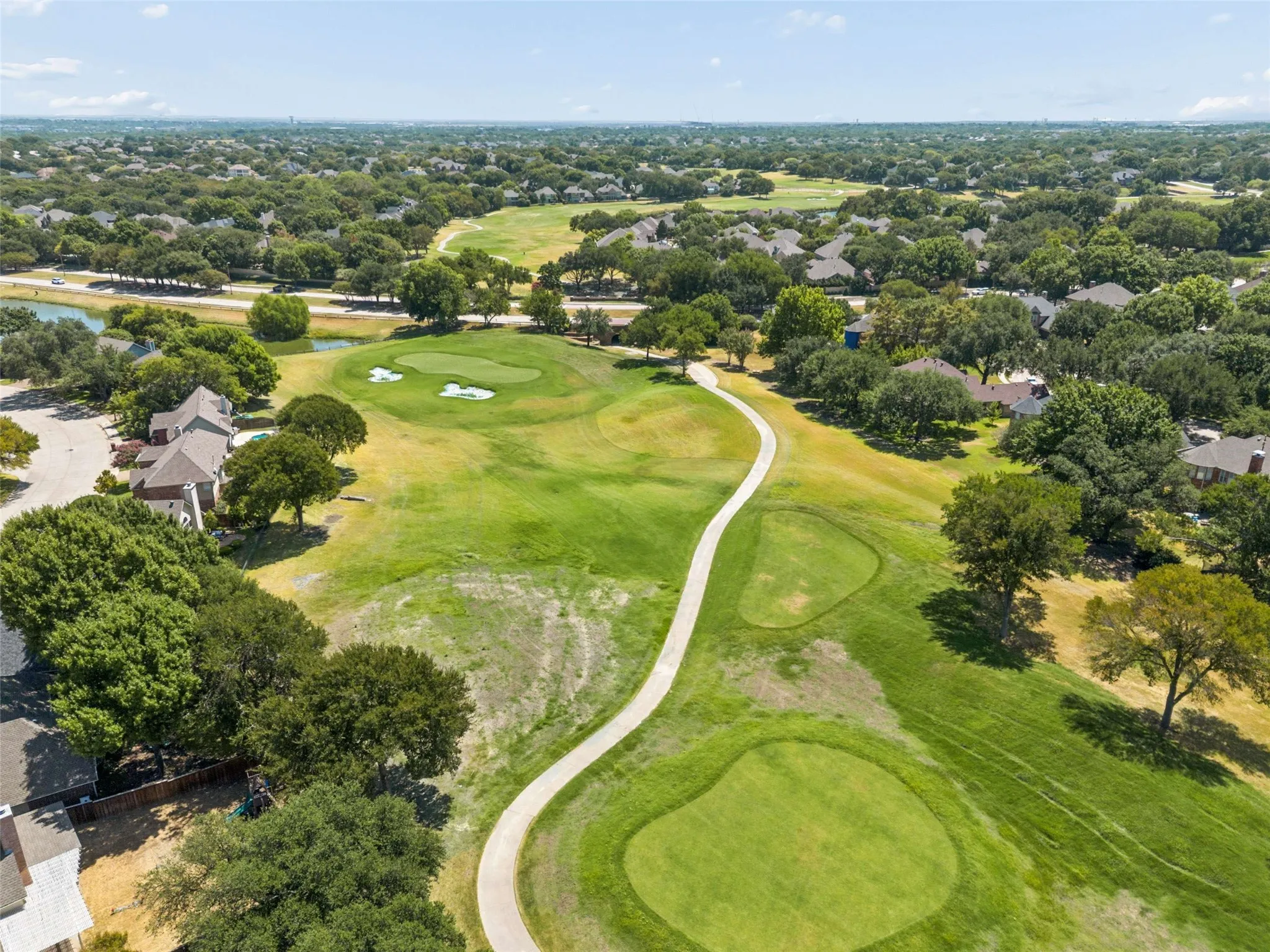 Expansive golf course setting featuring manicured greens, a paved cart path, surrounding mature trees, and nearby residential properties