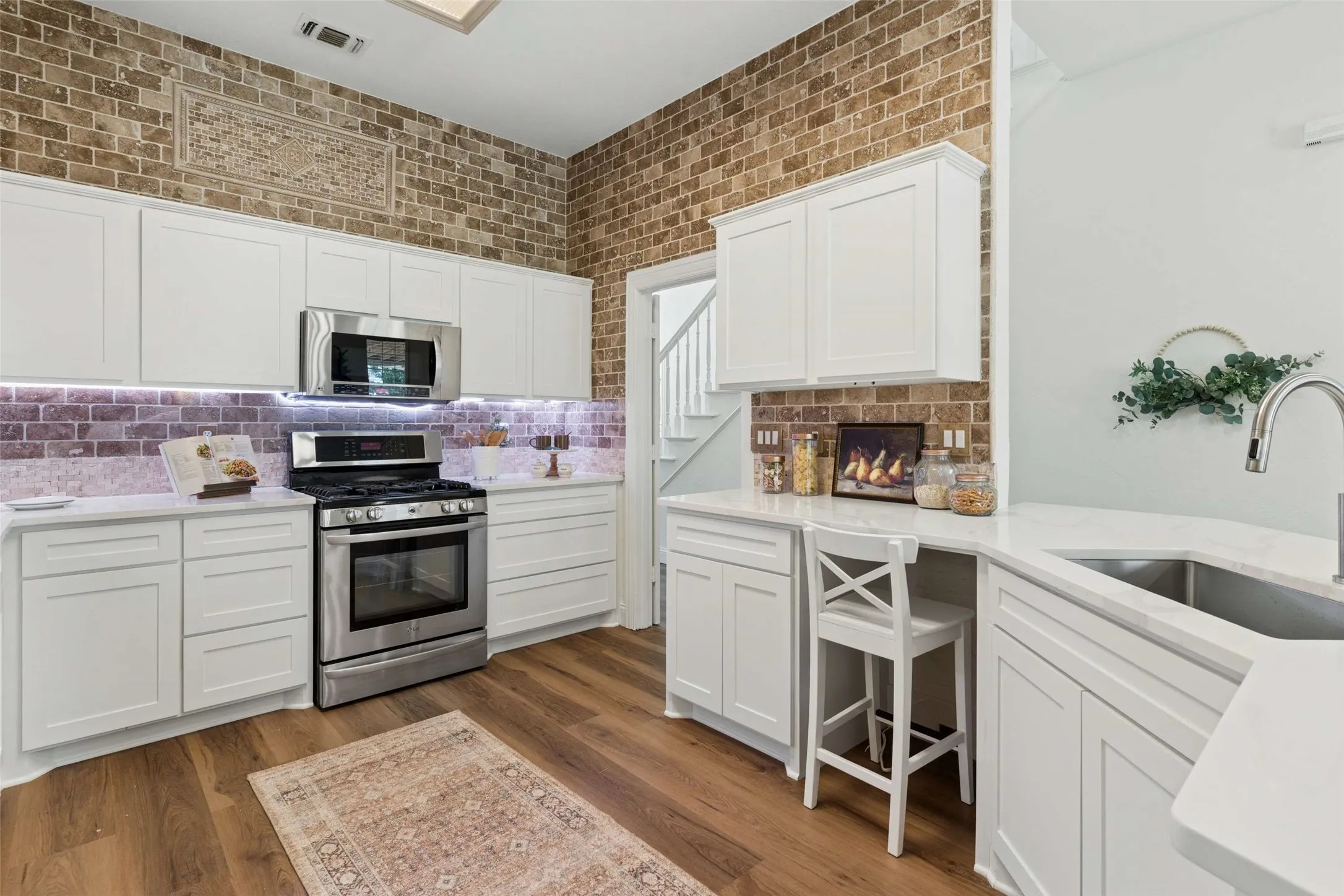 Wood-finish flooring with white cabinetry, stainless steel appliances, and brick-look wall accents