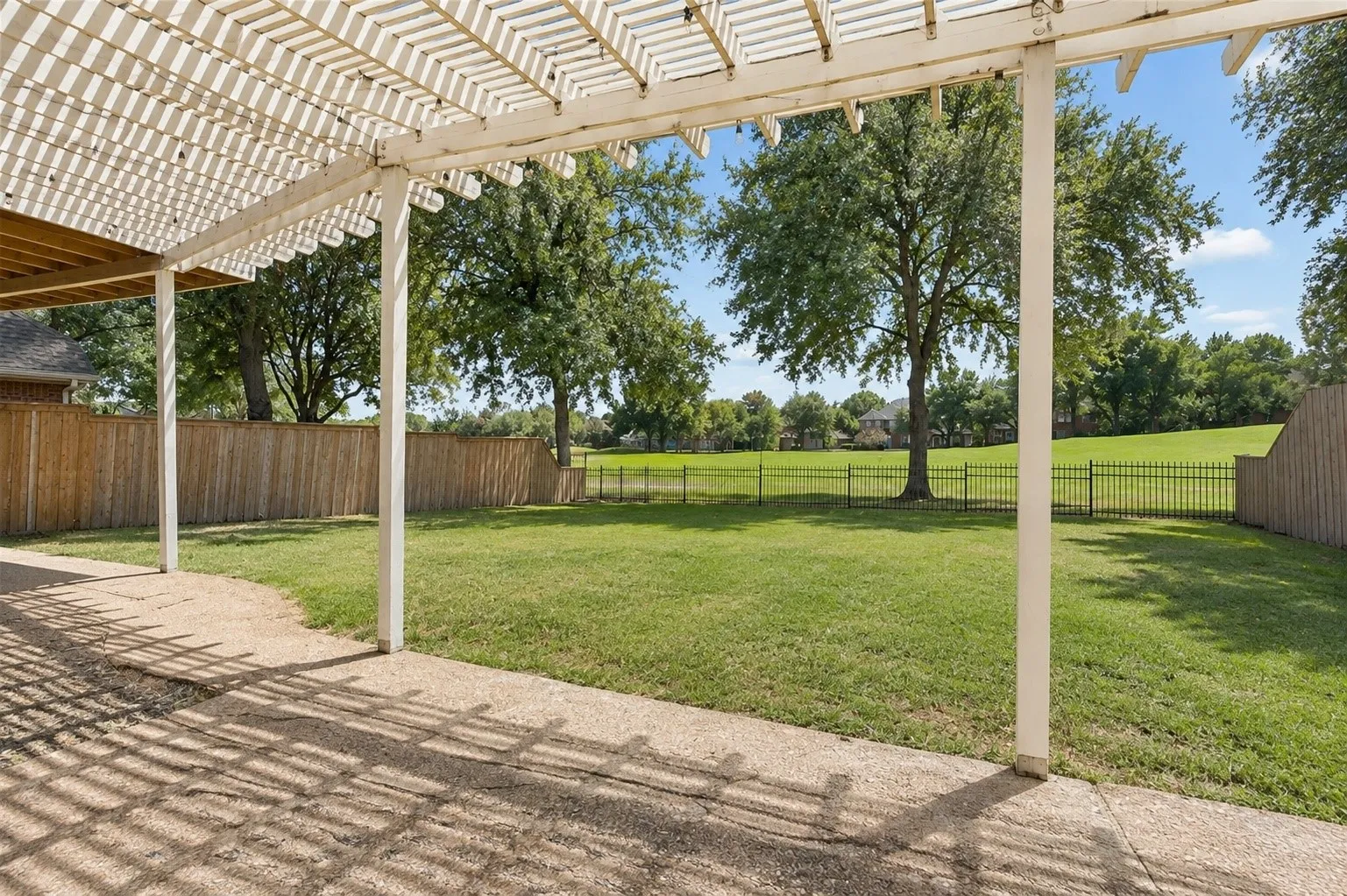 Pergola-covered patio with concrete flooring, overlooking a green lawn with a black metal fence and mature trees