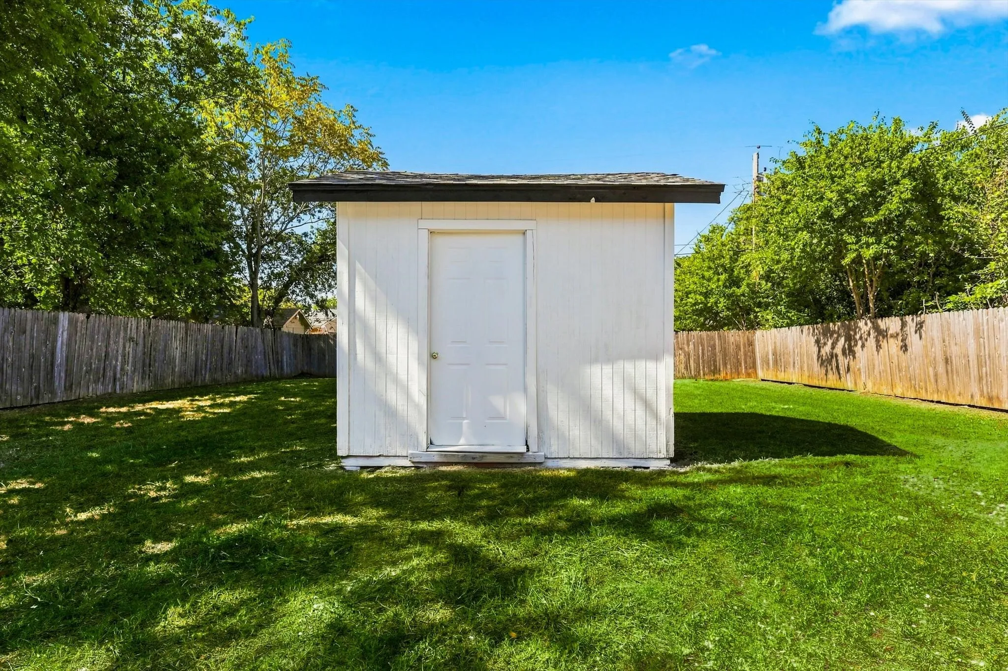 Detached storage shed with a panel door, set on a grassy lawn, bordered by a wooden fence