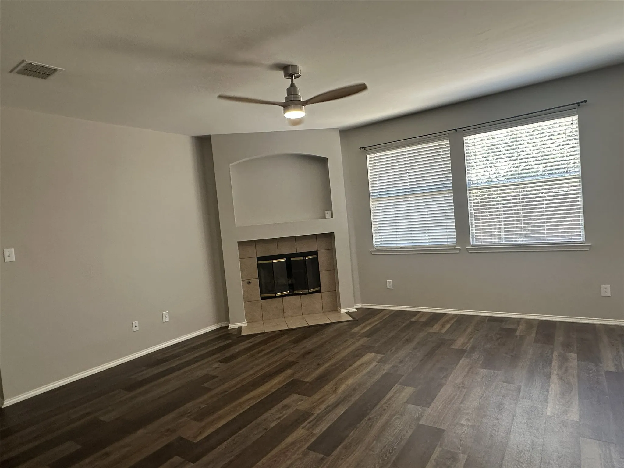 Wood-finish flooring, corner fireplace with tile surround, built-in niche, ceiling fan, and multiple windows with blinds