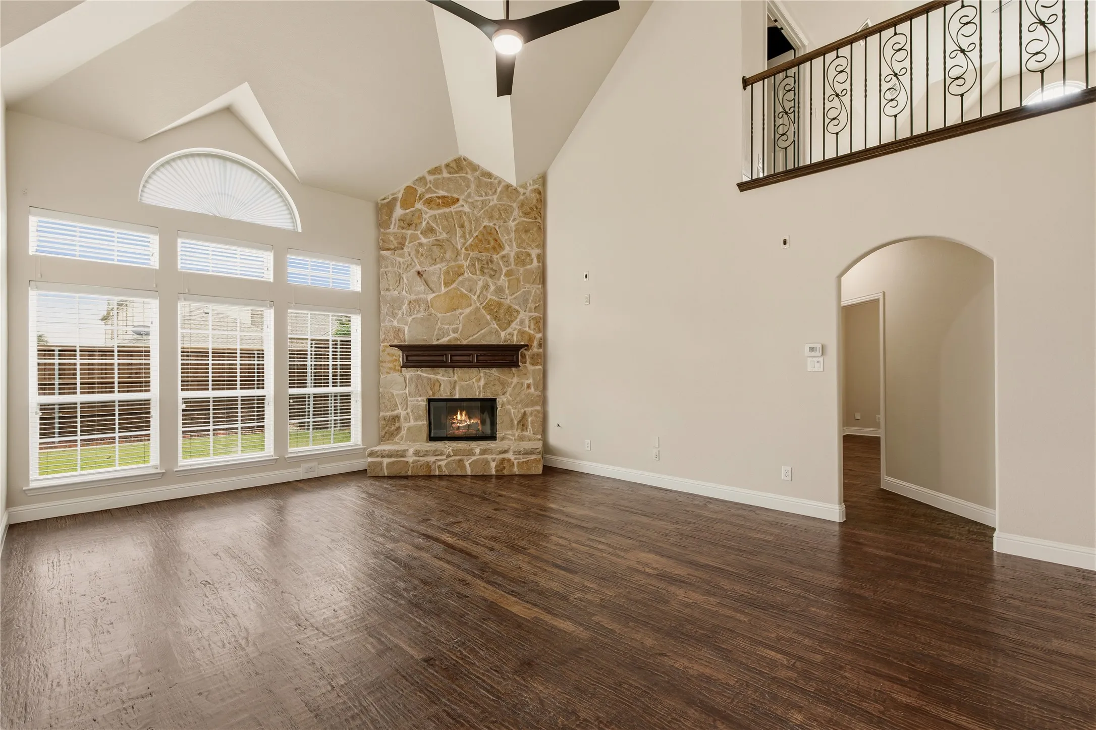Spacious living area featuring high vaulted ceilings, expansive windows with an arched transom, a stone fireplace with a wood mantle, wood-finish flooring, and a second-story overlook with an ornate iron railing