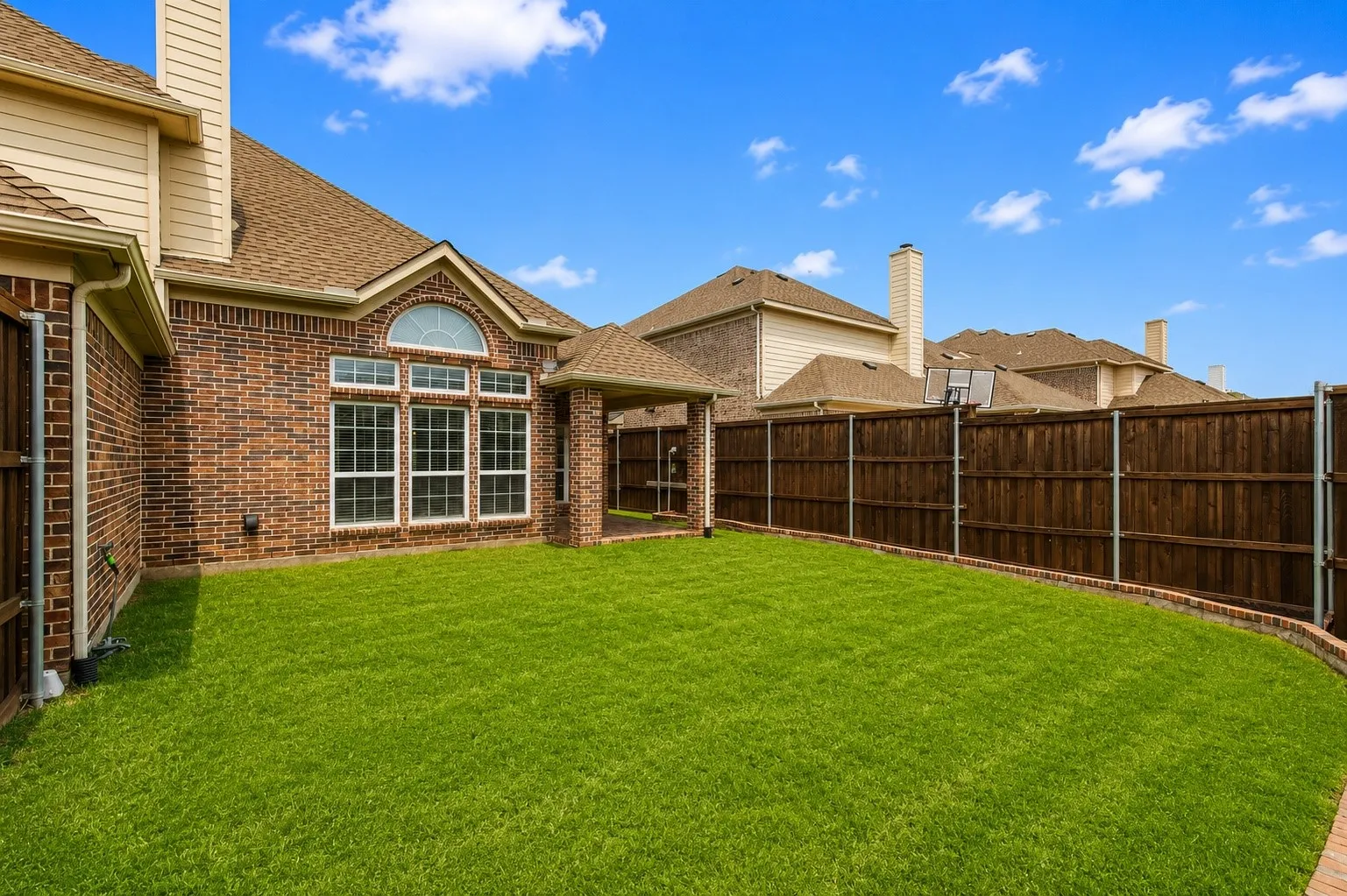 Spacious fenced backyard with a manicured lawn, brick perimeter, and covered patio
