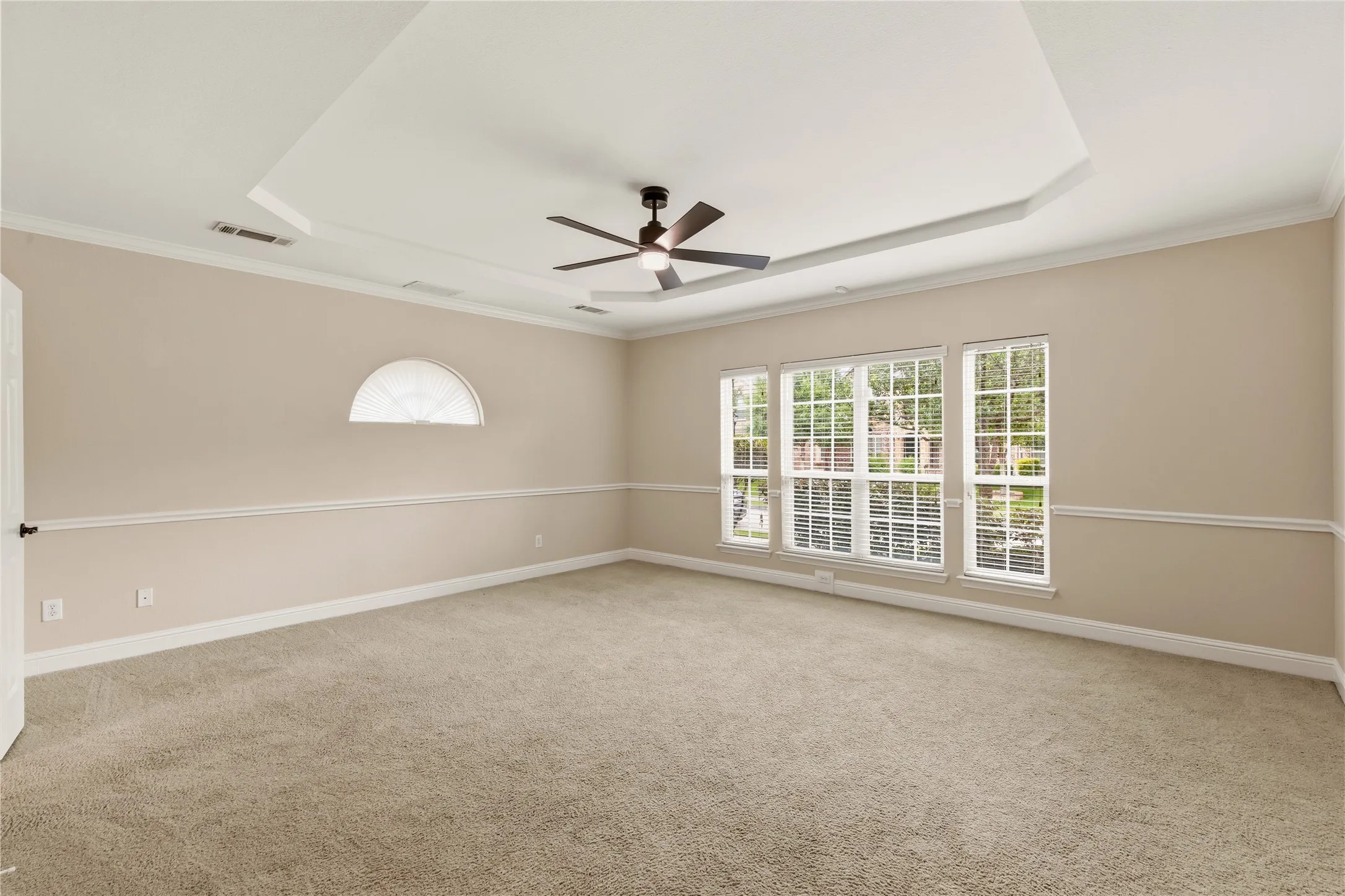 Spacious room featuring a tray ceiling with a ceiling fan, three large windows with white blinds, a half-round transom window, light-colored carpeting, and a chair rail molding