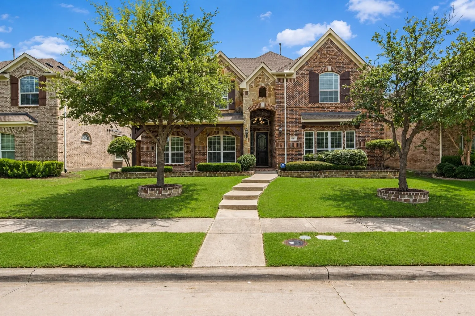 Brick and stone facade residence featuring a covered entry, established landscaping, decorative window shutters, a concrete sidewalk, and a manicured lawn