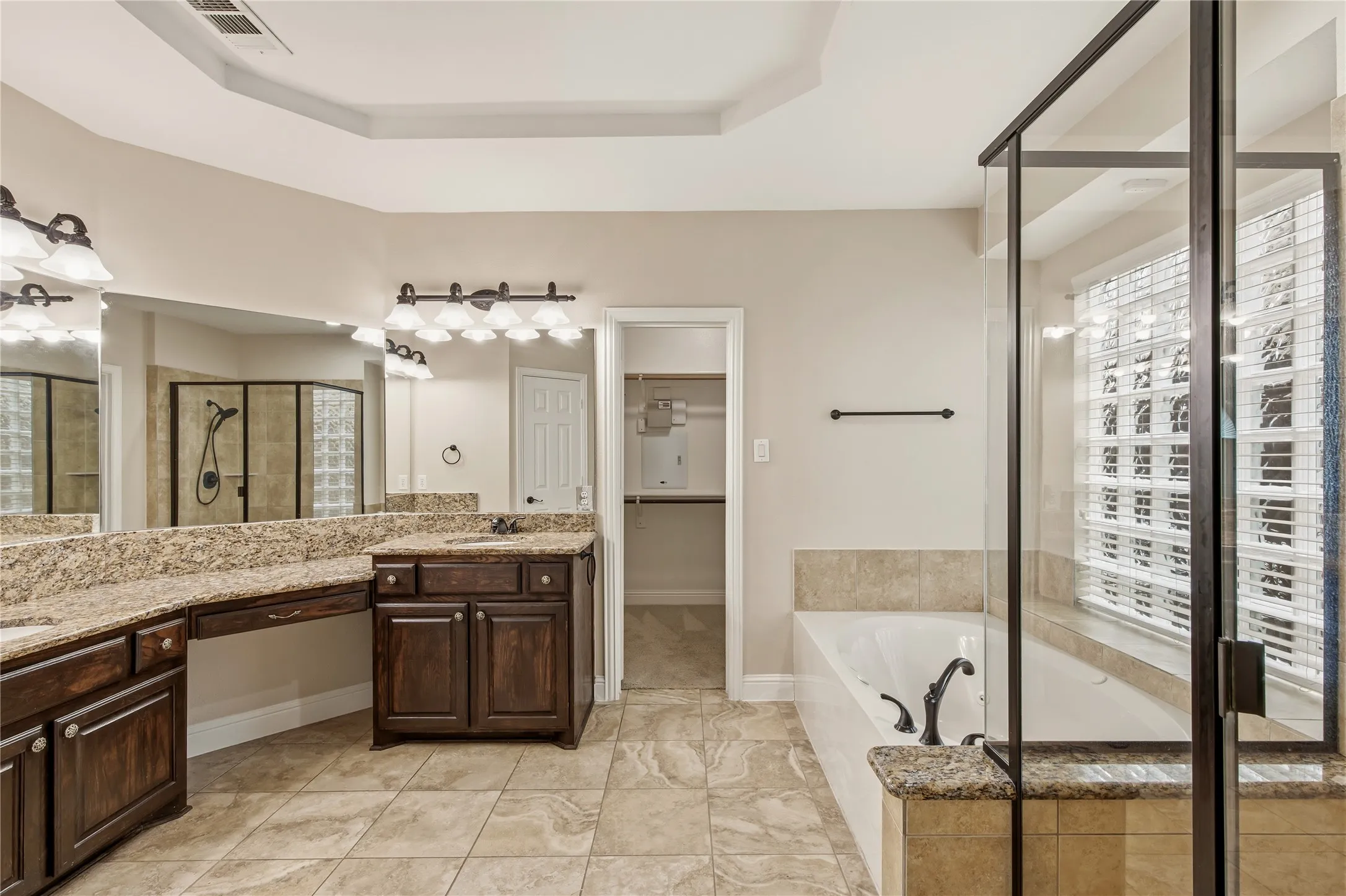 Bathroom featuring a dual vanity with dark wood cabinetry and granite countertops, a built-in soaking tub, a separate glass-enclosed shower with tile surround, and a tray ceiling