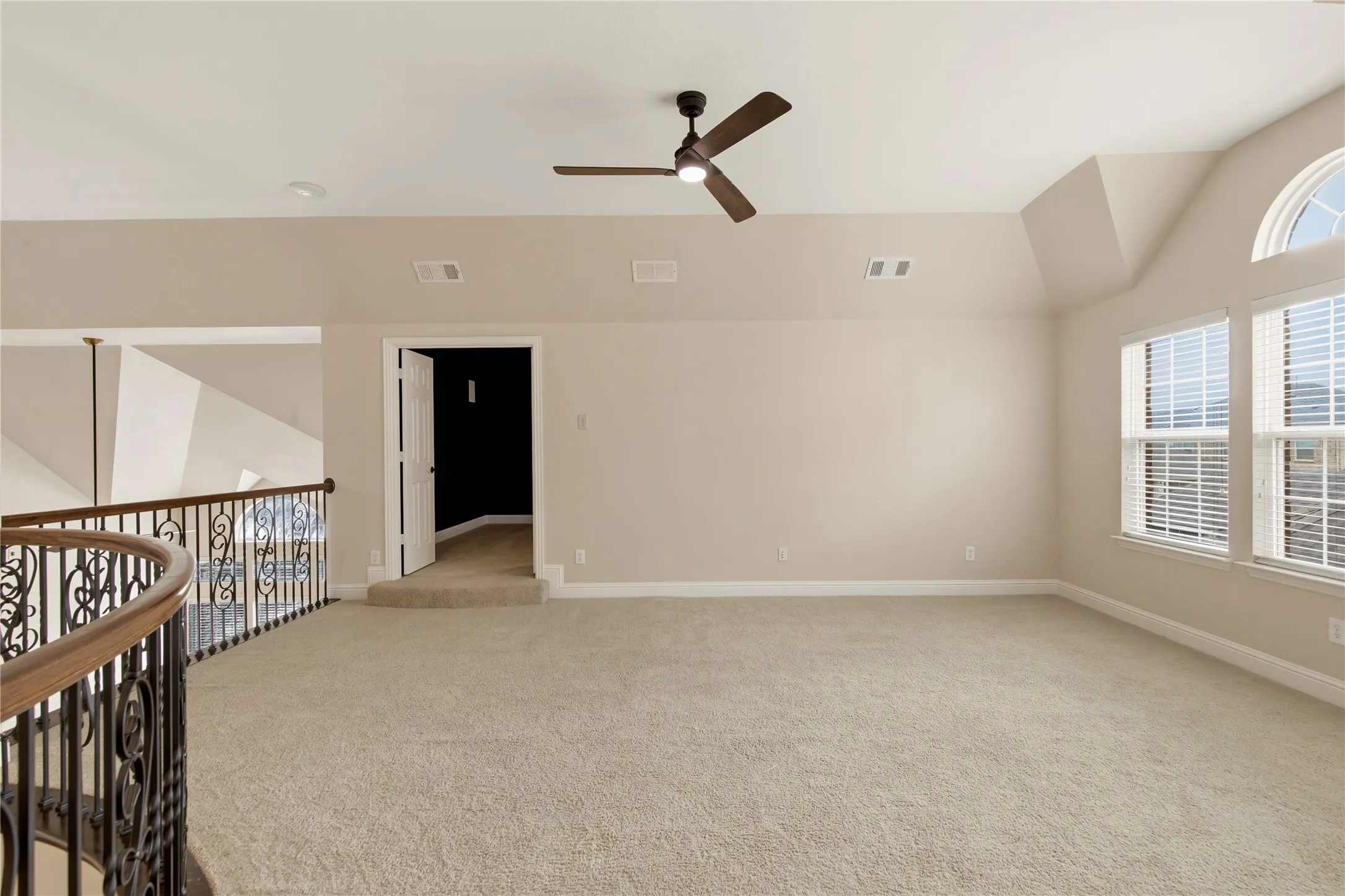 Spacious carpeted room featuring a ceiling fan, multiple windows with blinds, an arched window, and a decorative wrought iron and wood-finish railing overlooking a lower level