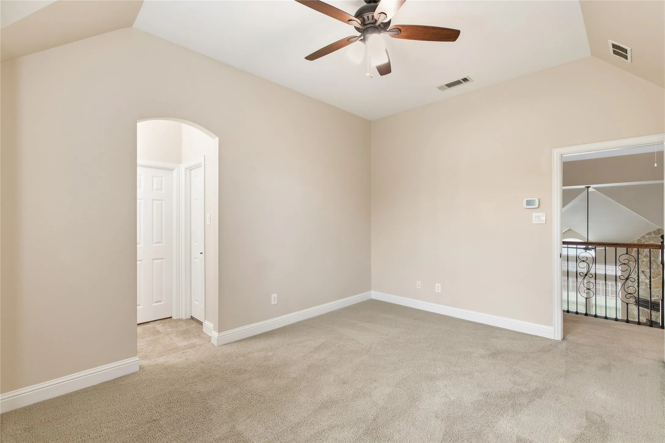 Carpeted room featuring a vaulted ceiling with a ceiling fan, an arched entryway, white baseboards, and a doorway opening to a wrought iron railing