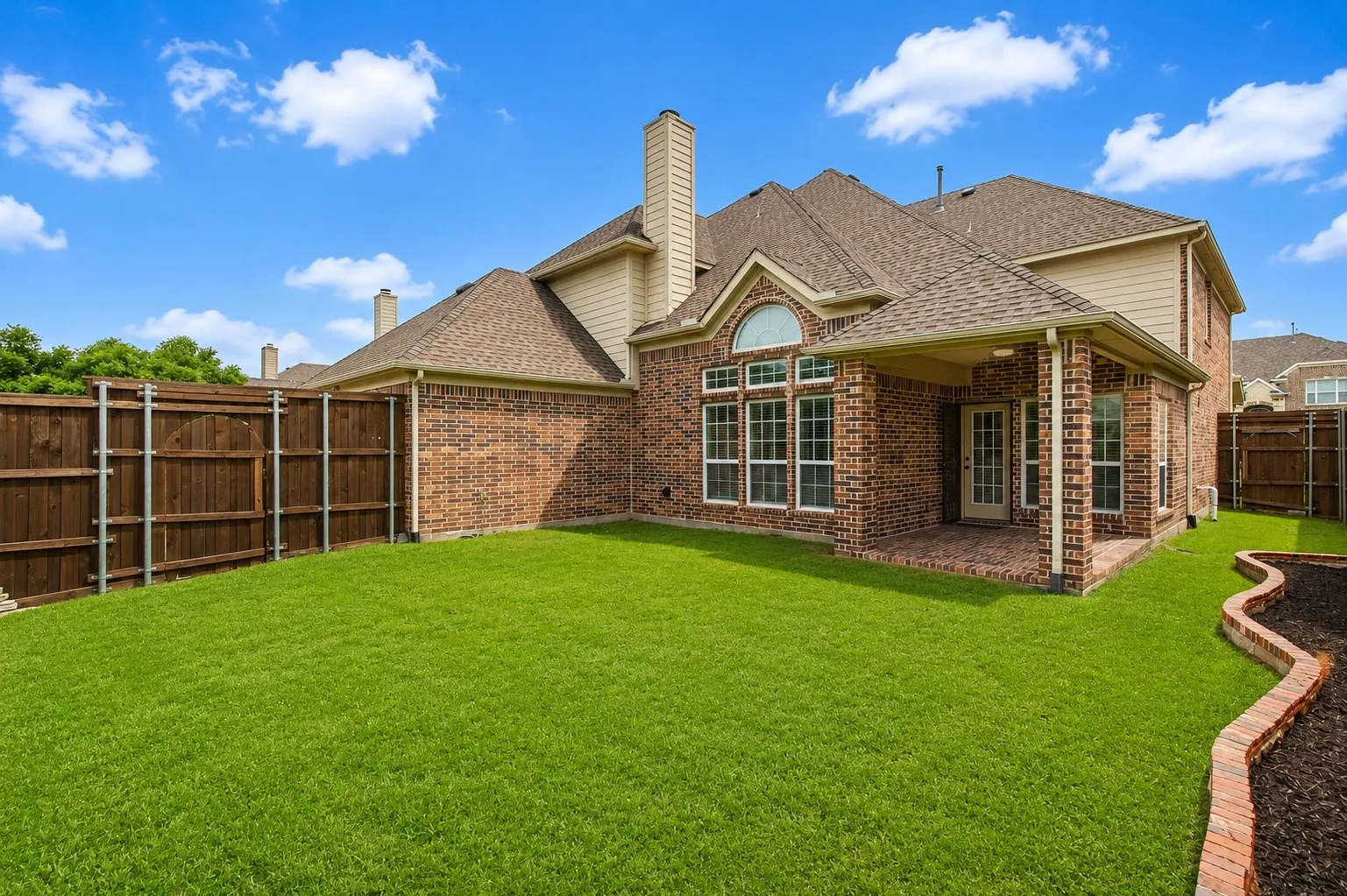 Spacious backyard featuring a covered brick patio, wood fencing, and a curved brick planter