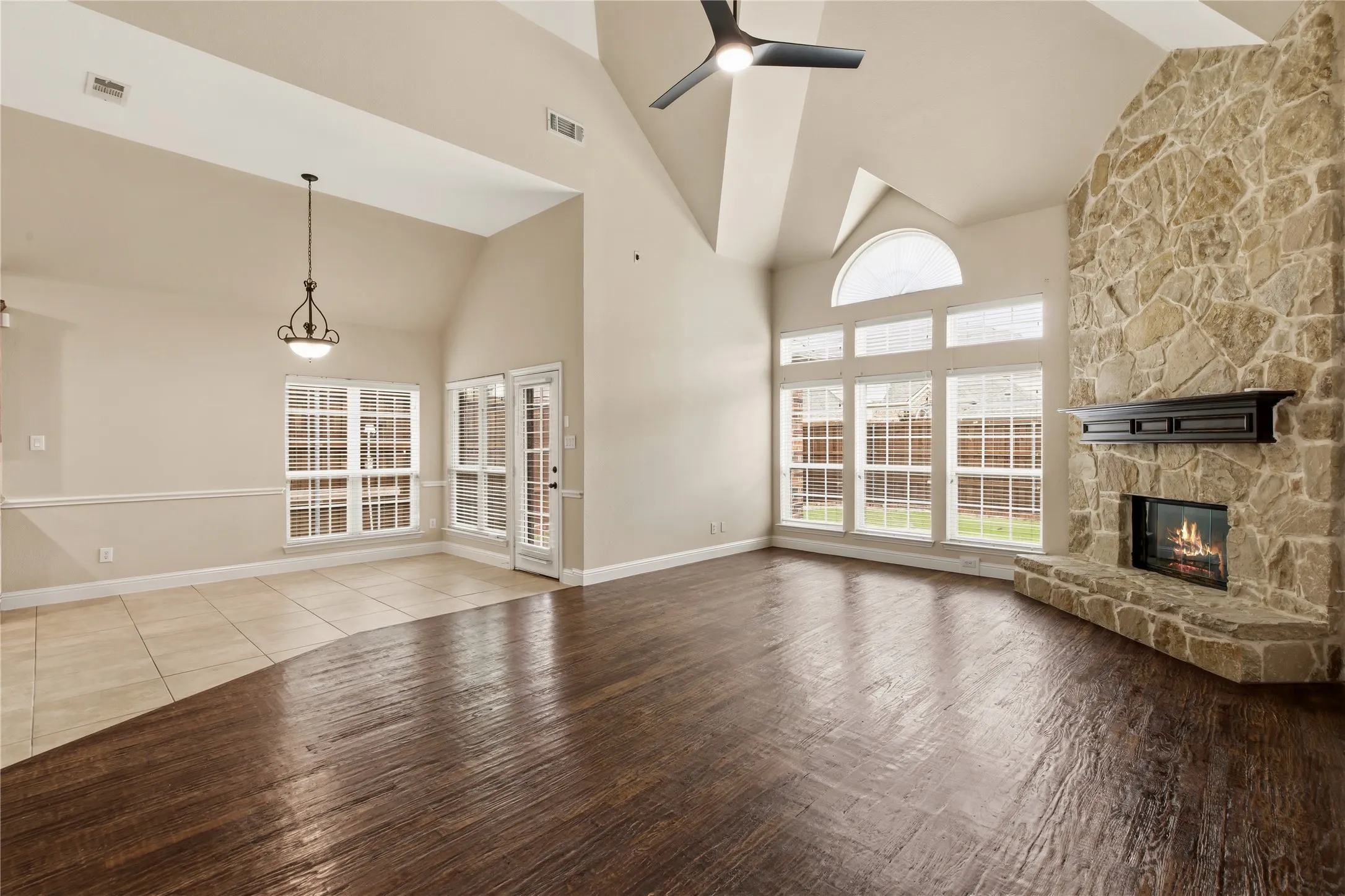 Spacious living area featuring a vaulted ceiling, stone-clad fireplace with mantel, dark wood-finish flooring, and large windows