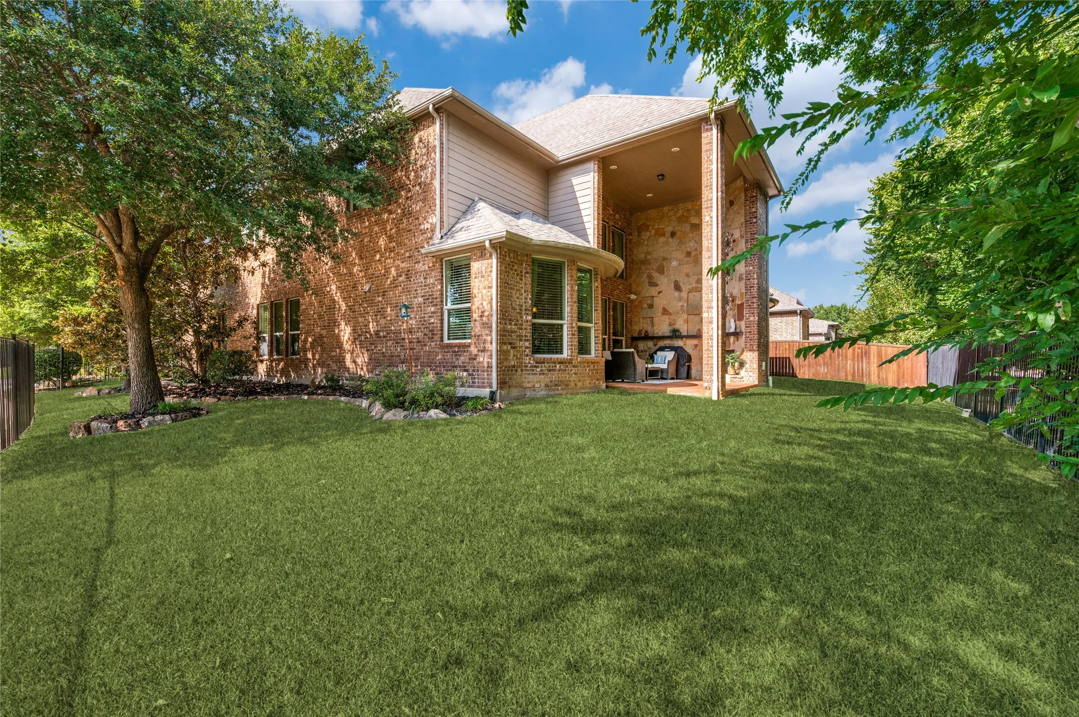 This lush backyard features a well-manicured lawn, mature trees, and professional stone-edge landscaping. The space includes a covered patio equipped with a stone fireplace, perfect for outdoor entertaining.