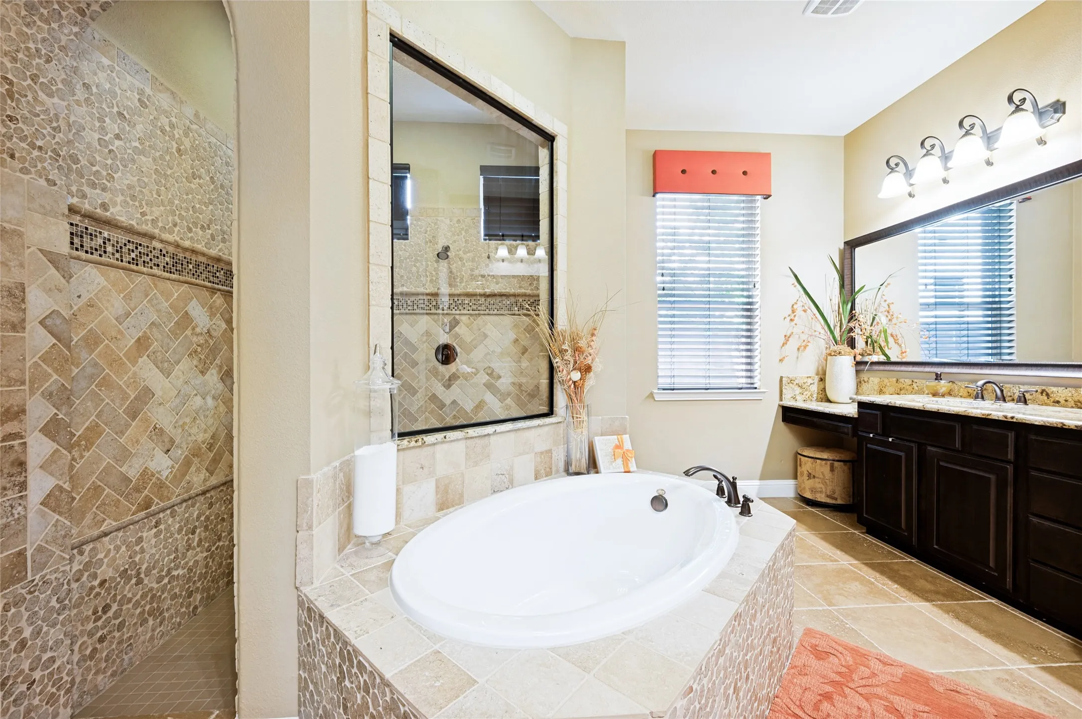 This primary bathroom features natural light, a large soaking tub with custom tile surround, a walk-in double shower accented by pebble and herringbone tile work, dual vanities including a dedicated makeup station, a linen closet, and expansive framed mirrors.