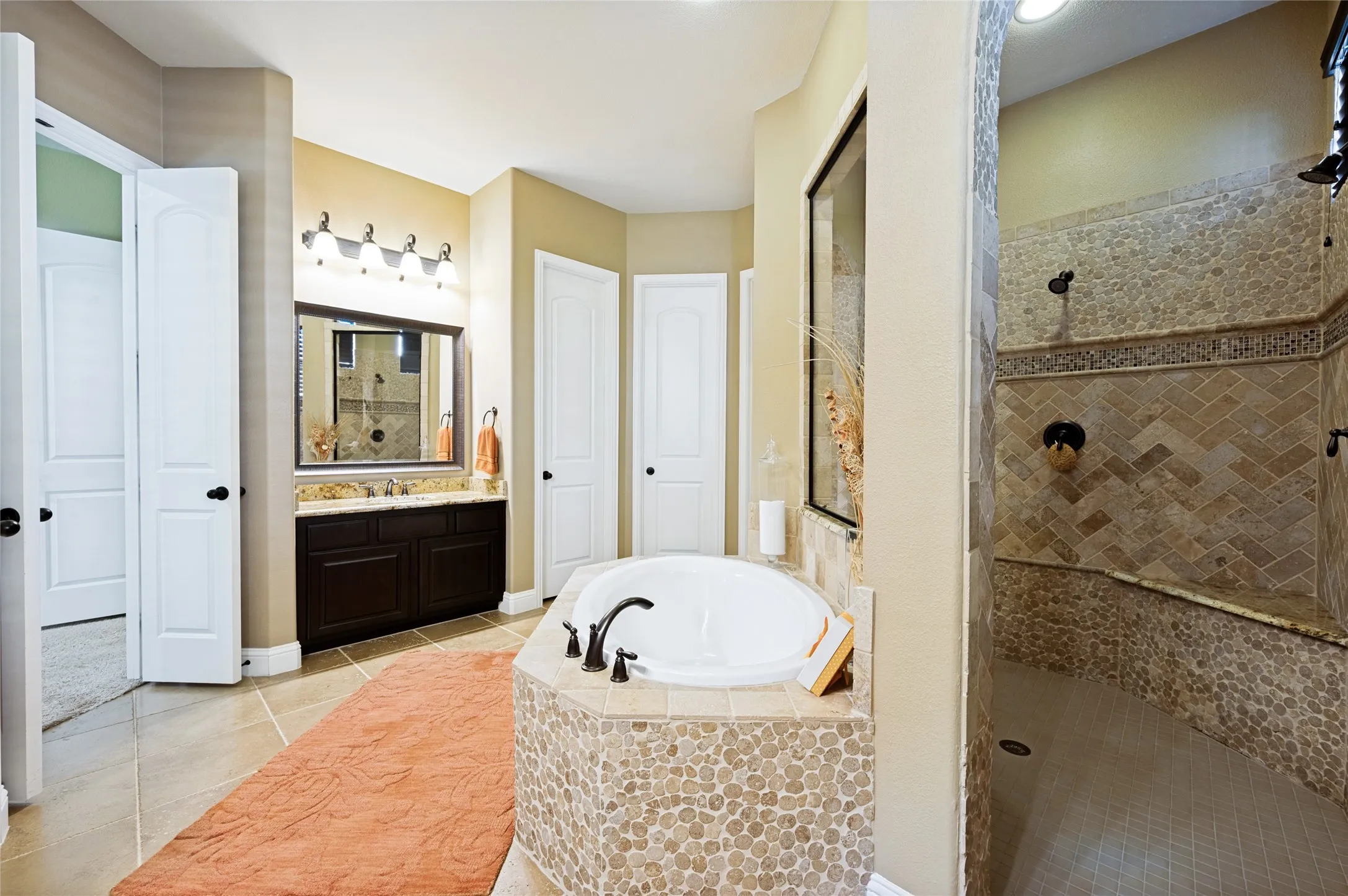 This luxurious primary bathroom includes an oval soaking tub with a stone tile surround, stone tile flooring, and separate vanity areas featuring granite countertops. Additionally, the space offers a double walk-in shower finished in stone tile, complemented by dual walk-in closets.