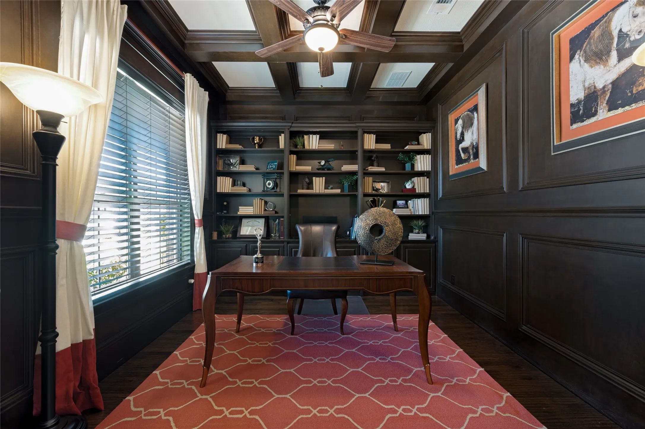 This home office features refined custom wood paneling, integrated built-in shelving, hand-scraped hardwood flooring, a coffered ceiling, and a ceiling fan with integrated lighting.