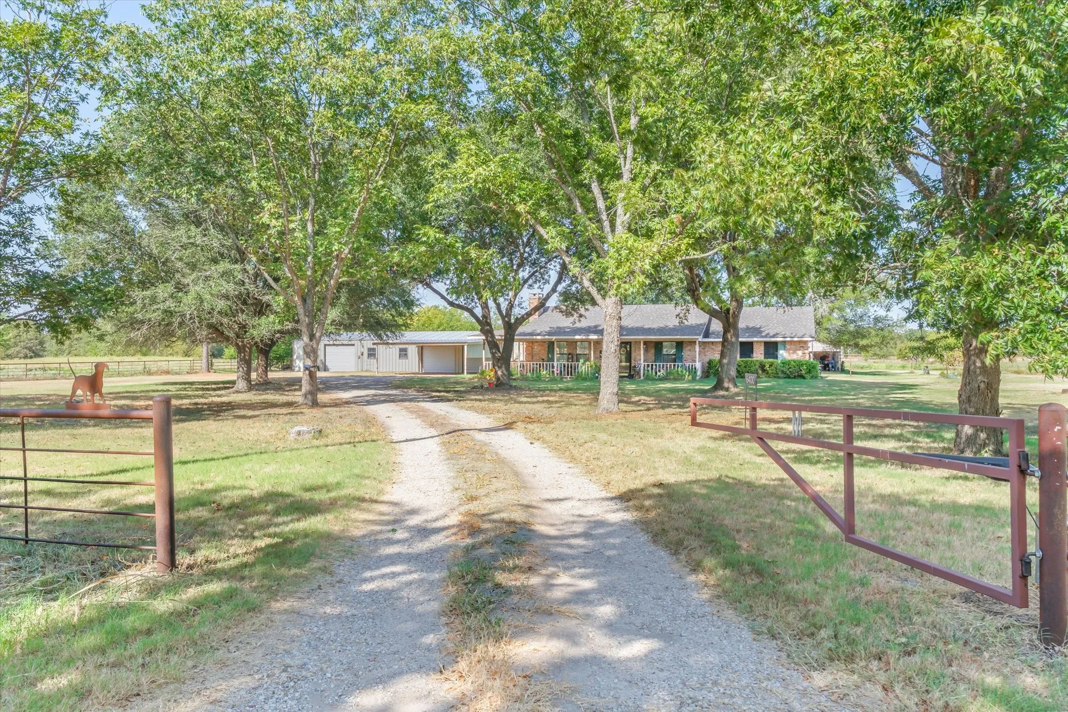 Gravel driveway with metal gate, mature trees, and a ranch-style house with an extended porch
