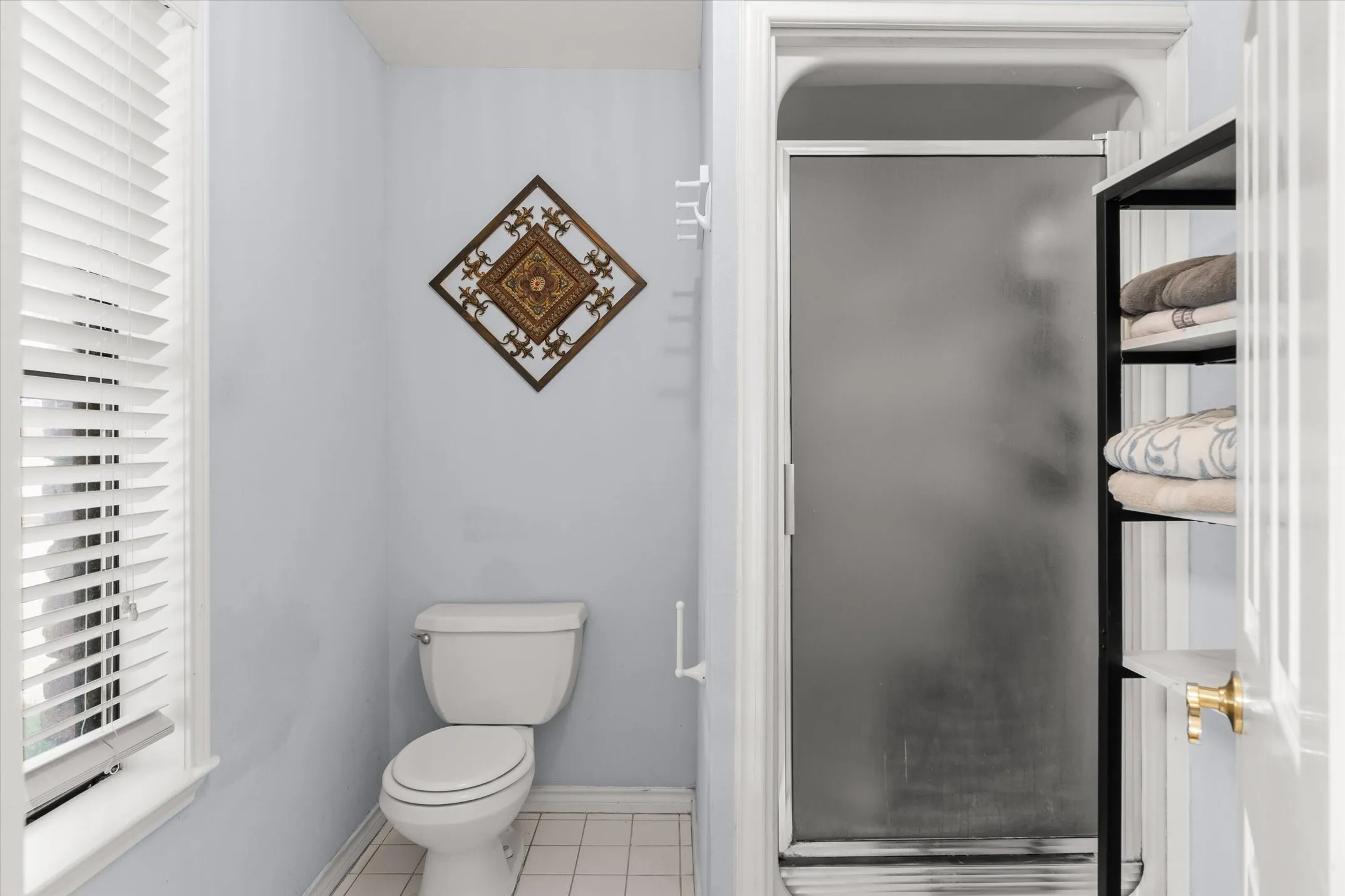 Window with white louvered blinds, white toilet, tiled flooring, built-in shelving, and a shower with a frosted glass enclosure