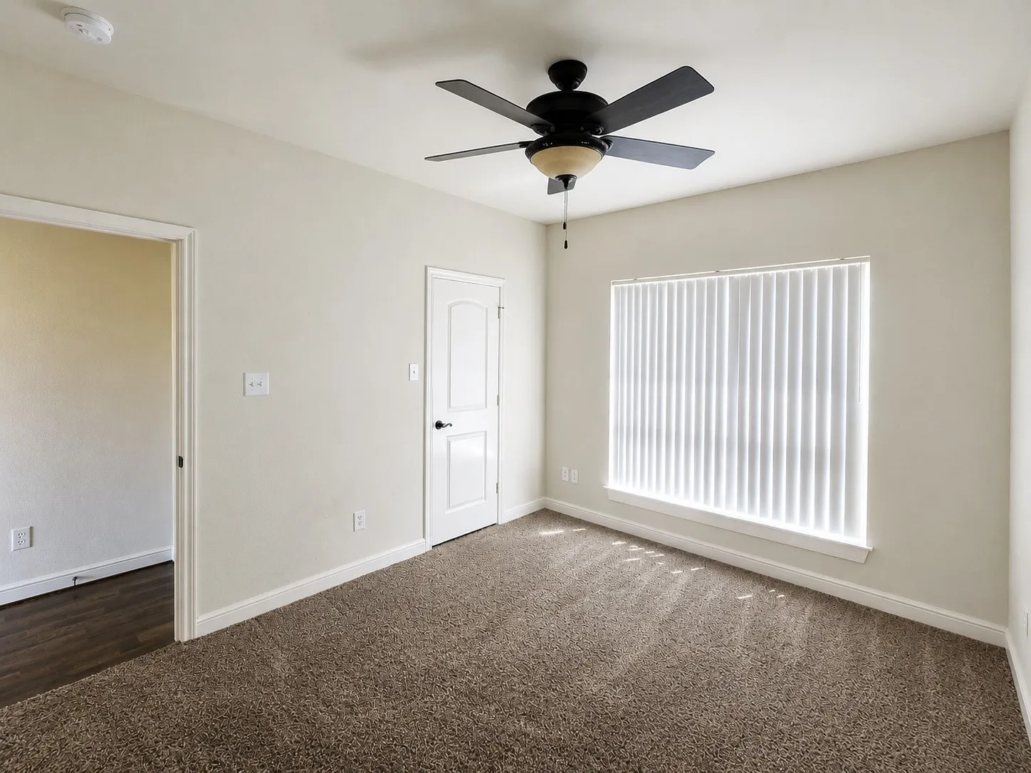 Textured carpet flooring, ceiling fan with integrated lighting, window with vertical blinds, painted interior doors with dark hardware, and light-toned wall finishes