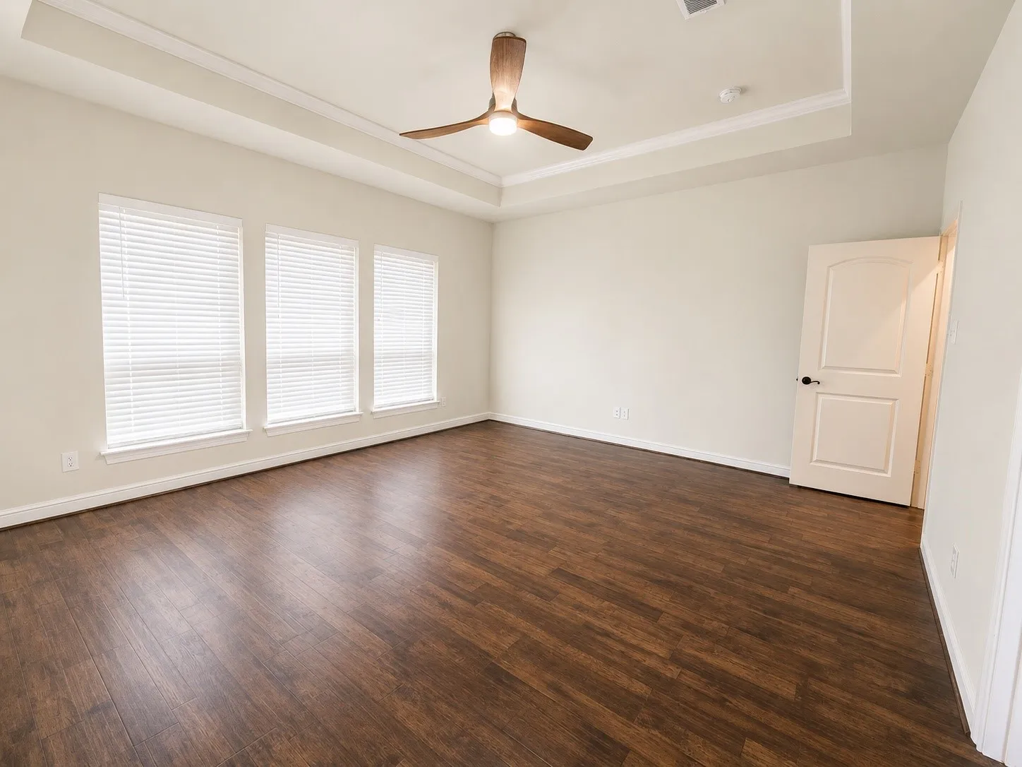 Tray ceiling with crown molding, ceiling fan, three windows with blinds, wood-finish flooring, and panel door