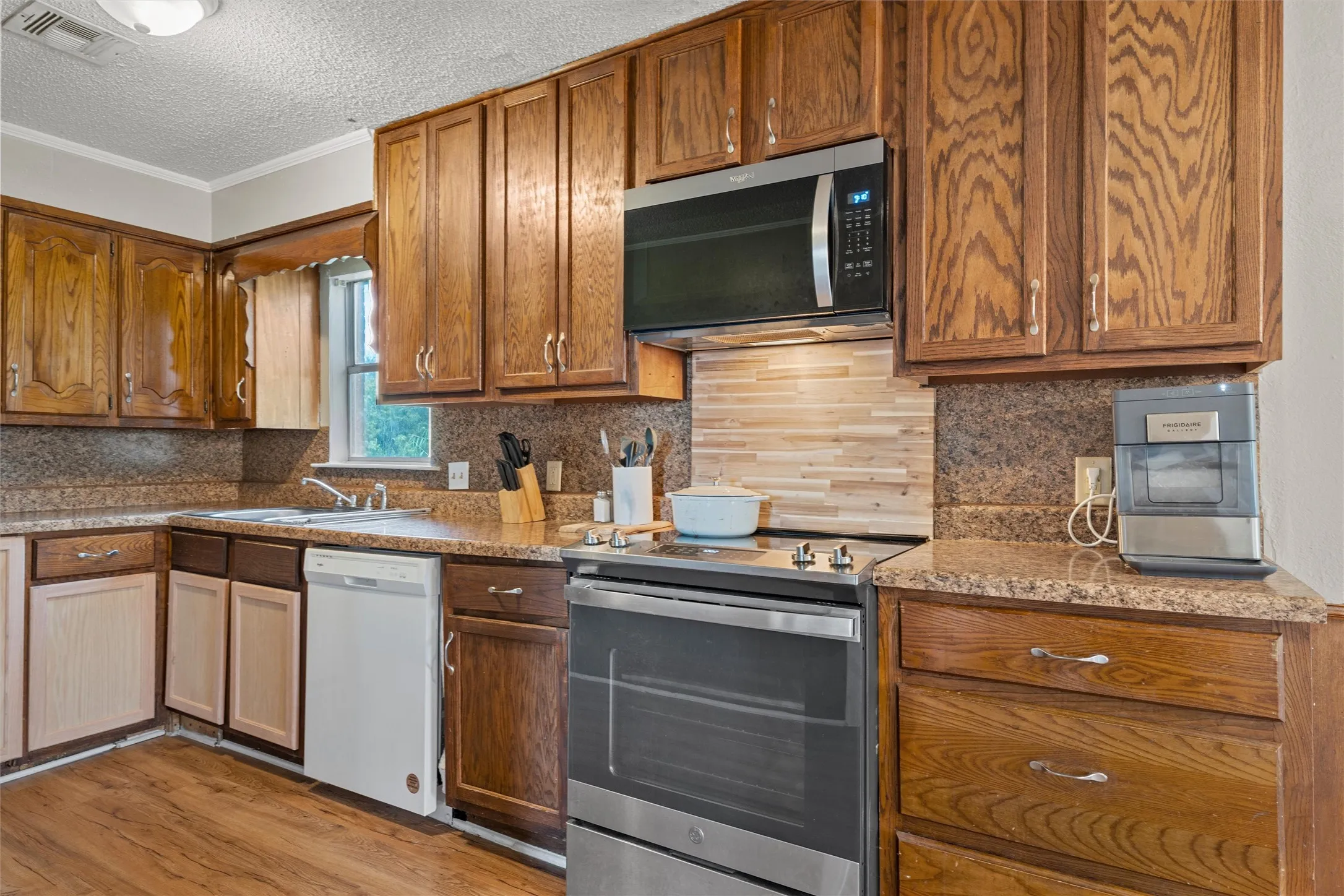 Wood-finish flooring, wood cabinetry with hardware, built-in stainless steel microwave, stainless steel range, and a dual-basin sink with a window