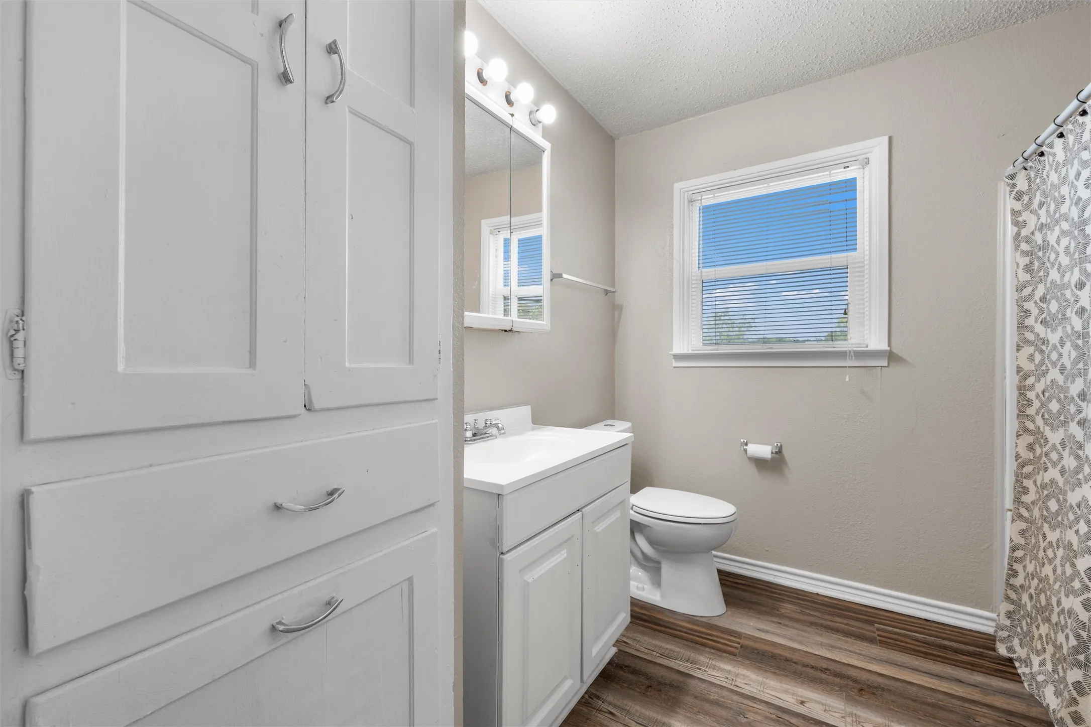 Wood-finish flooring, white vanity with integrated sink, built-in storage cabinetry, vanity mirror with overhead lighting, and a window with blinds