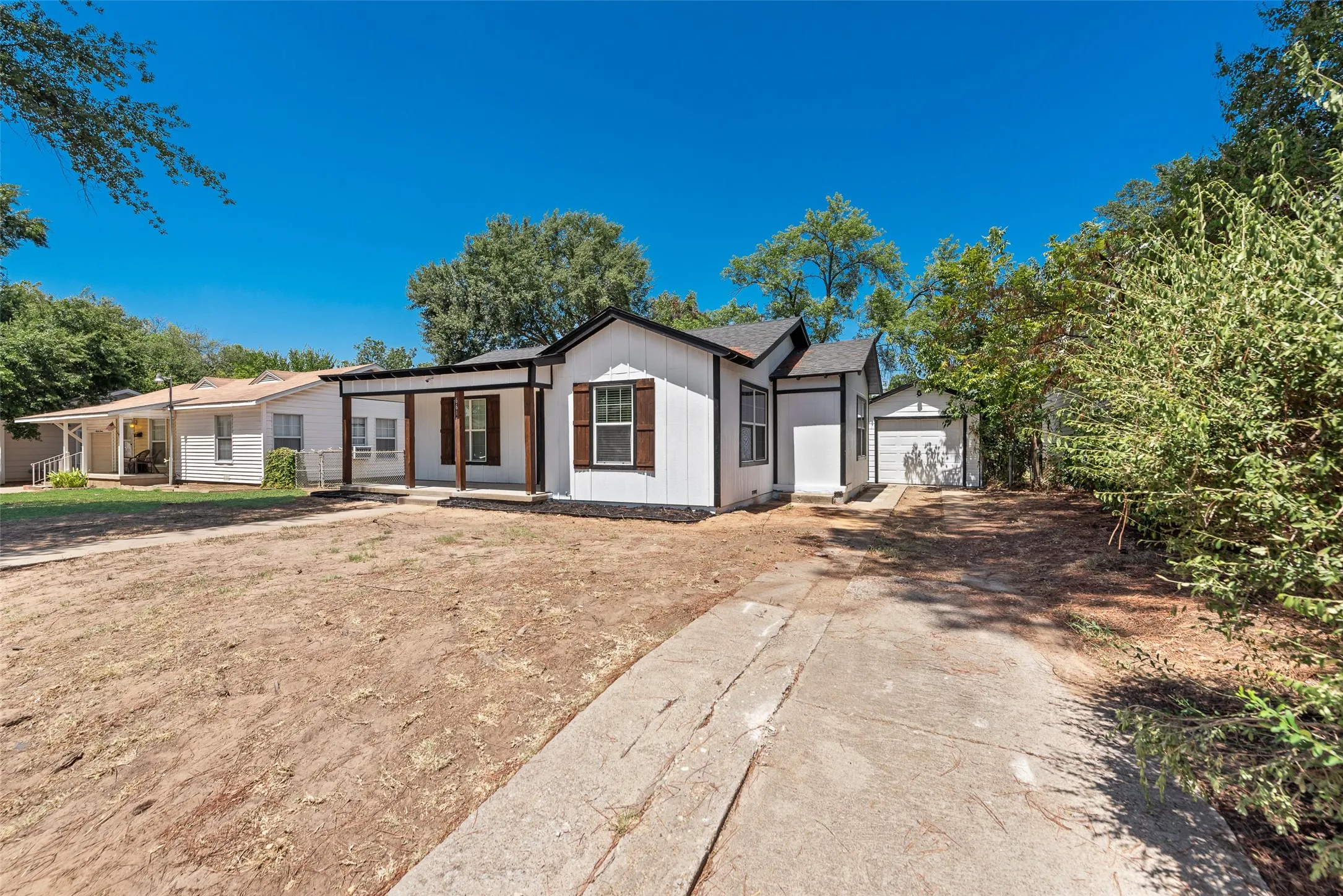 White exterior with dark trim and brown window shutters, covered front porch with wood posts, asphalt shingle roof, and a detached garage