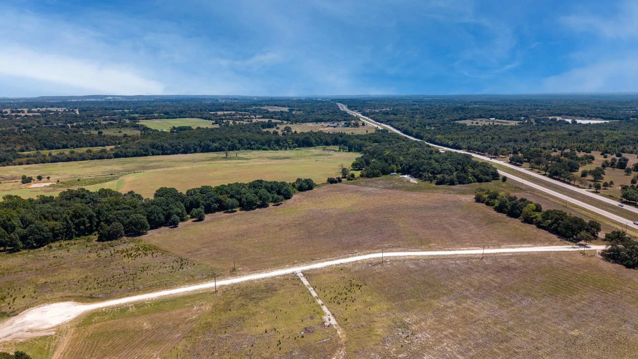 Expansive rural landscape featuring a highway, wooded areas, open fields, and a dirt road