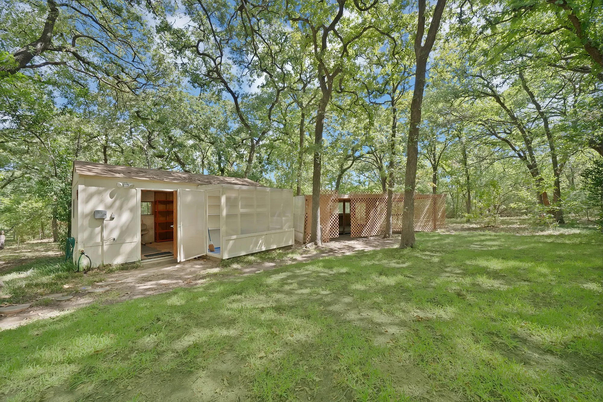 White-sided outbuilding with an open doorway, sheltered by mature trees, featuring an attached trellis structure