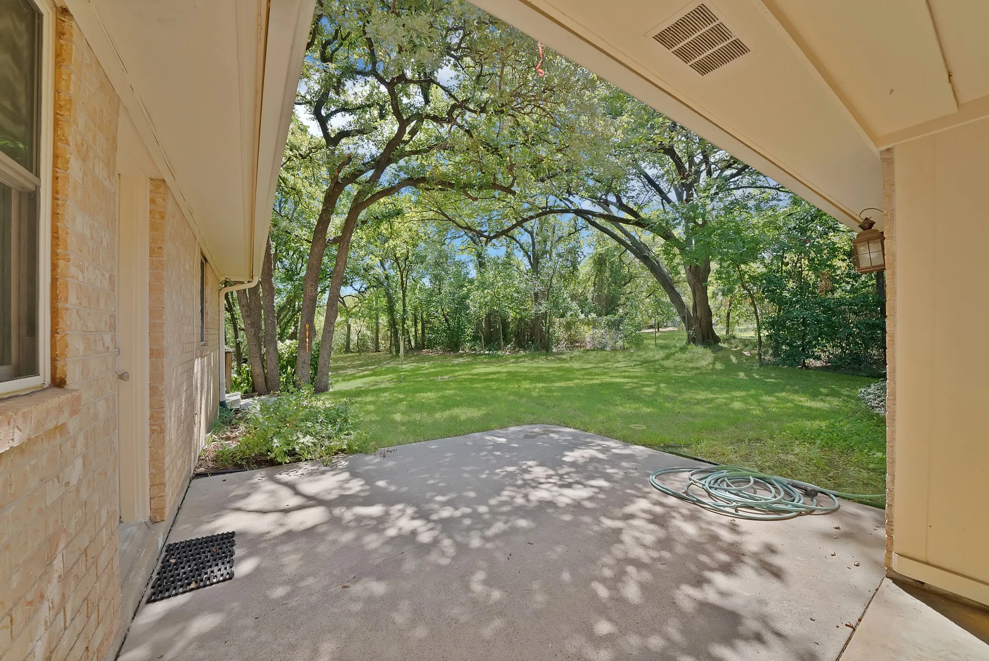 Covered patio with concrete flooring, brick exterior, and a large grassy area surrounded by mature trees