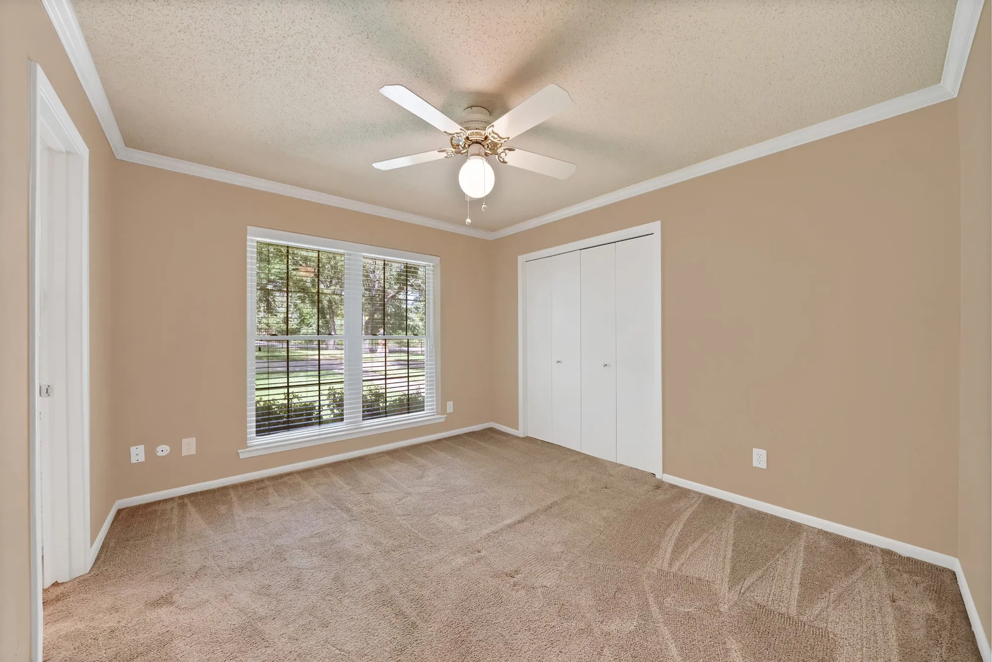 Neutral-toned carpeting, ample natural light from a large window, a ceiling fan with integrated lighting, and white bi-fold closet doors