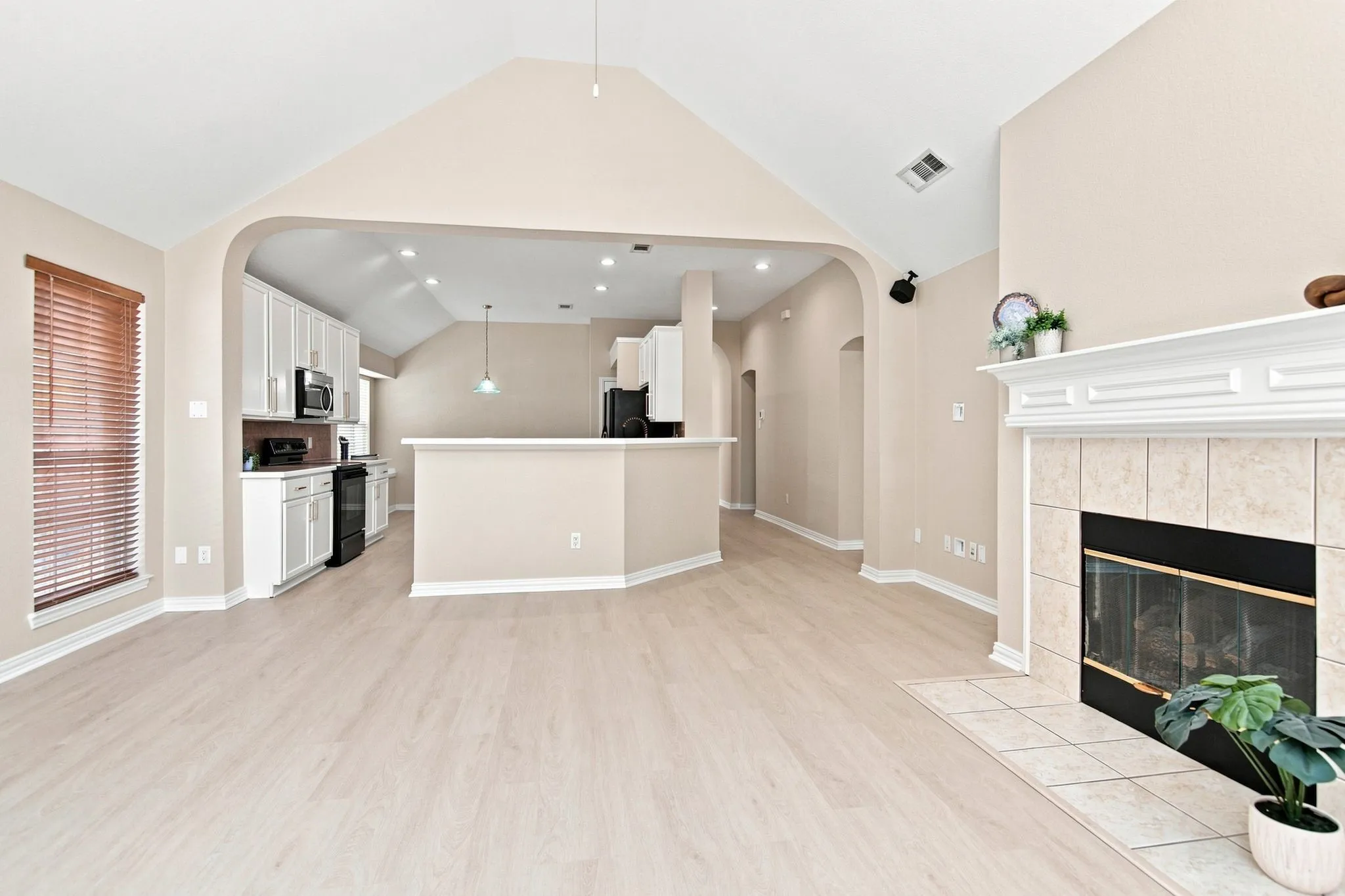 Vaulted ceiling, light wood-finish flooring, tiled fireplace with mantel, arched entryway, and white kitchen cabinetry with dark appliances