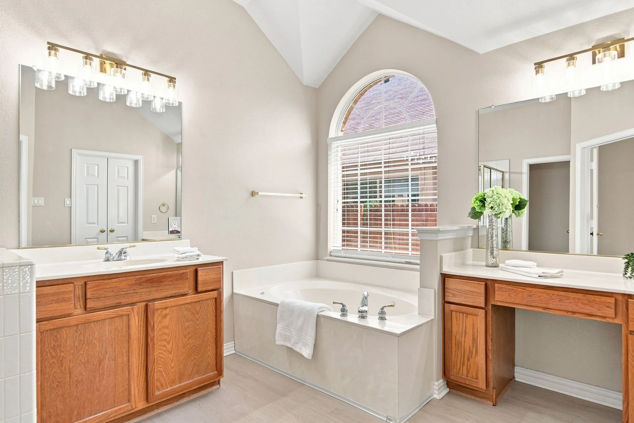 Arched window with blinds, dual vanity sinks, separate makeup vanity, built-in bathtub, and vaulted ceiling