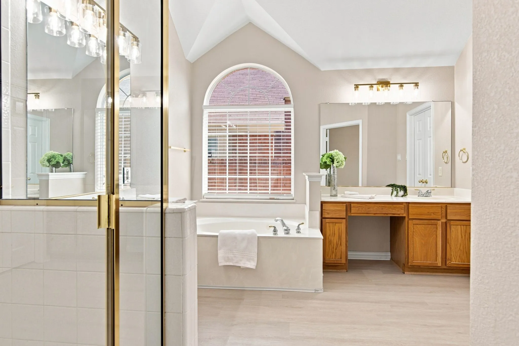 Vaulted ceiling, arched window with privacy blinds, built-in soaking tub, wood-finish vanity with dual sinks, and glass-enclosed shower with brass-finish framing
