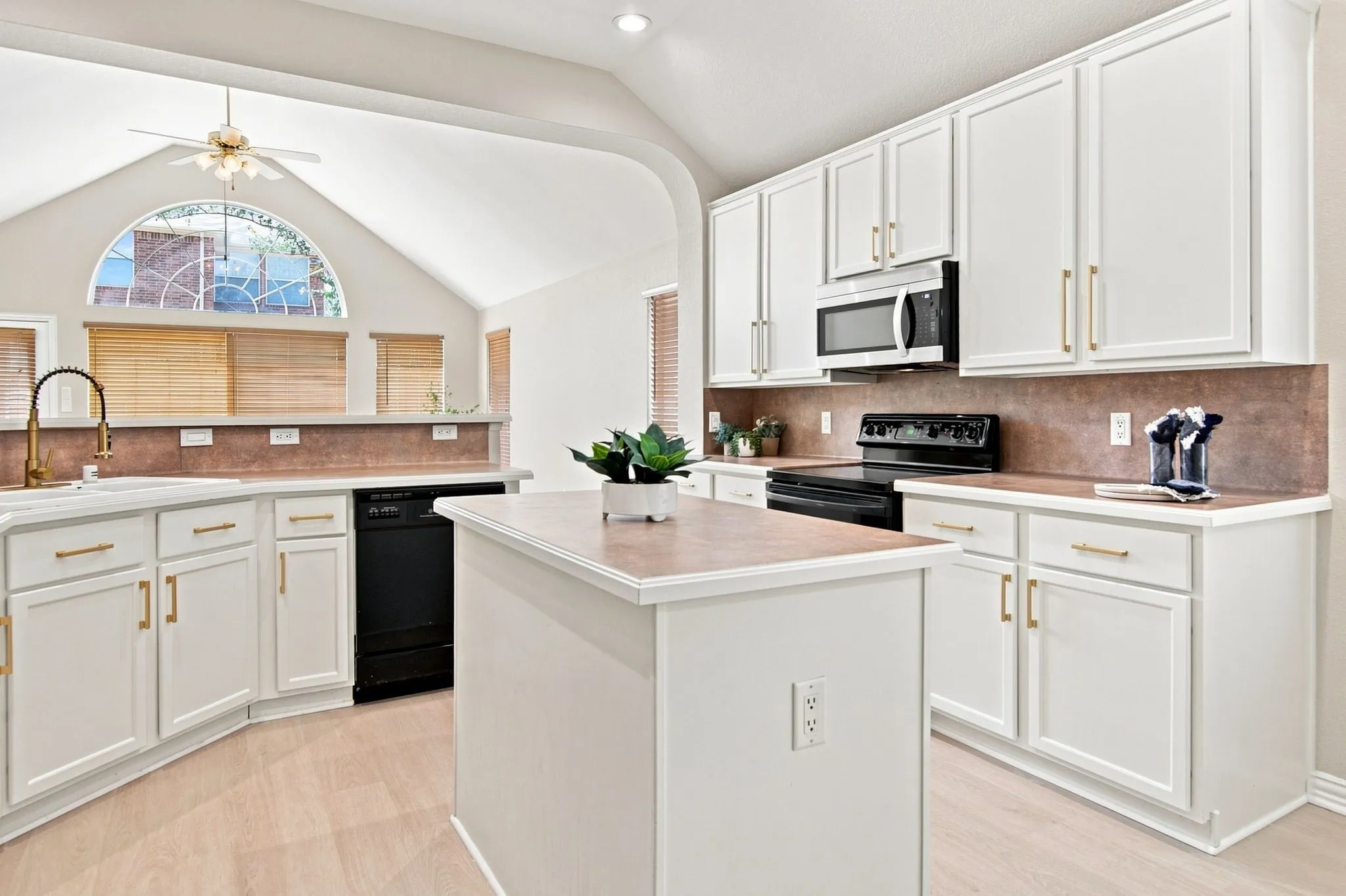 White cabinetry with gold-tone hardware, central kitchen island, black appliances, light wood-finish flooring, and vaulted ceiling with an arched window