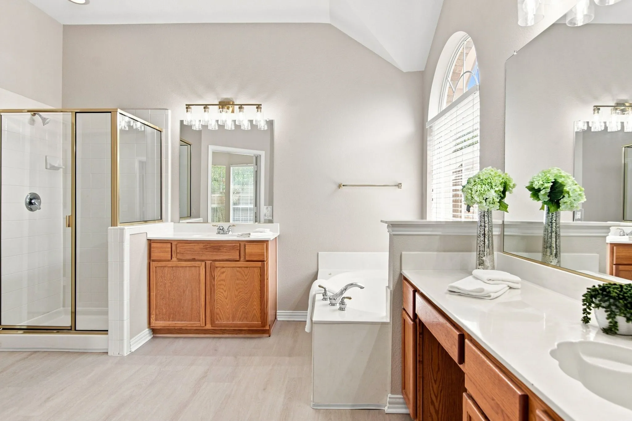 Dual wood-finish vanities with solid surface countertops, a built-in tub with deck-mounted fixtures, a glass-enclosed shower with tile surround, and an arched window