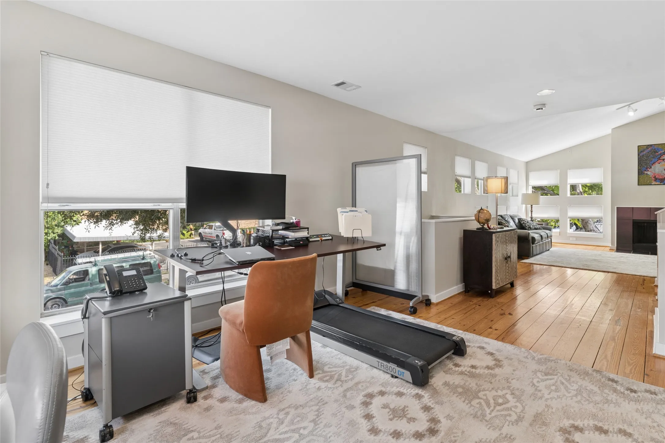 Bright home office featuring warm wood-finish flooring, expansive desk workspace with built-in storage, large windows with motorized roller shades, and abundant natural light