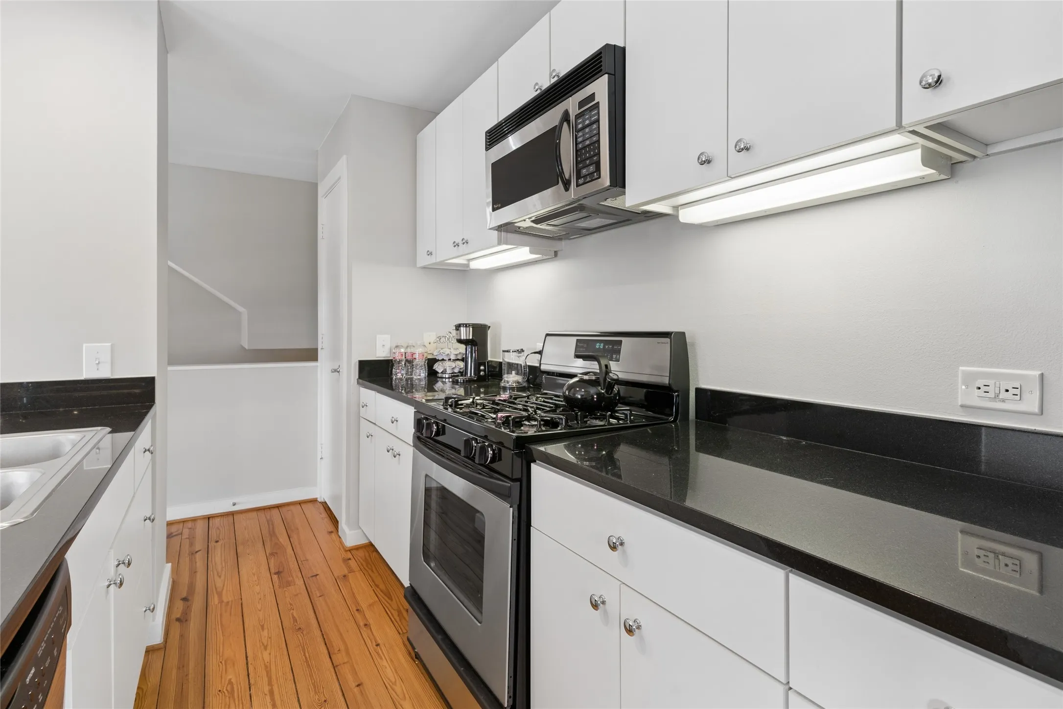 Galley kitchen featuring wood-finish flooring, white cabinetry, dark countertops, a stainless steel gas range, and an over-the-range microwave