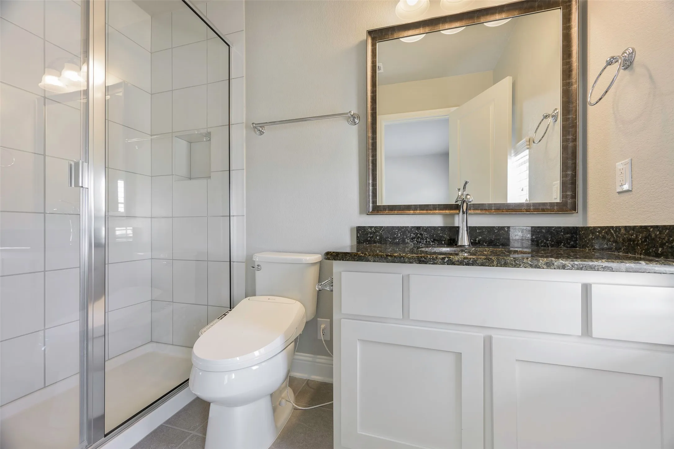 Glass-enclosed shower with white tiled walls, white shaker-style vanity, dark speckled countertop, and framed mirror