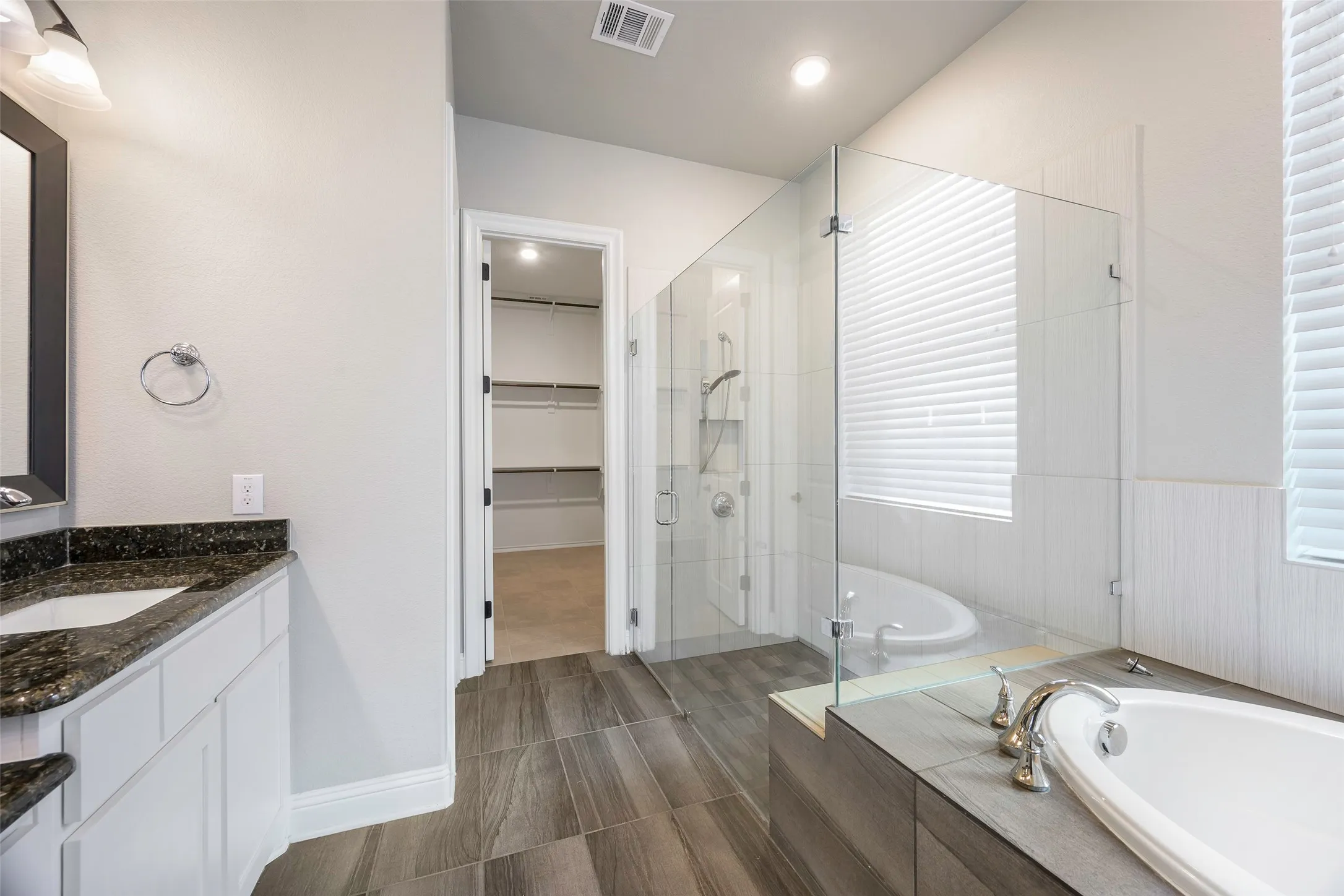 Dual vanity with dark stone countertop, separate walk-in shower with glass enclosure, freestanding soaking tub, and wood-finish flooring