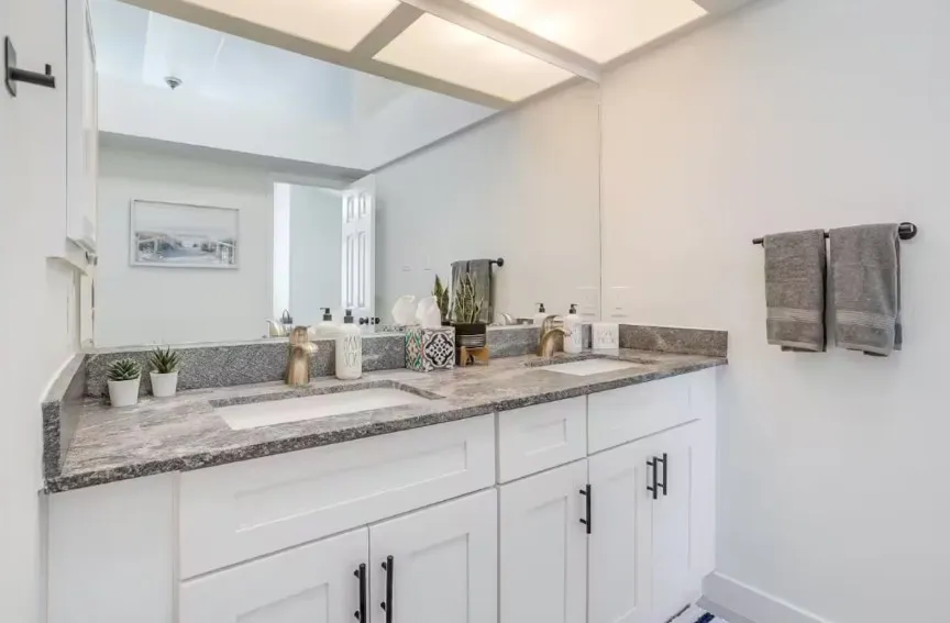 Dual vanity with undermount sinks, stone countertop with matching backsplash, and white cabinetry with matte black hardware