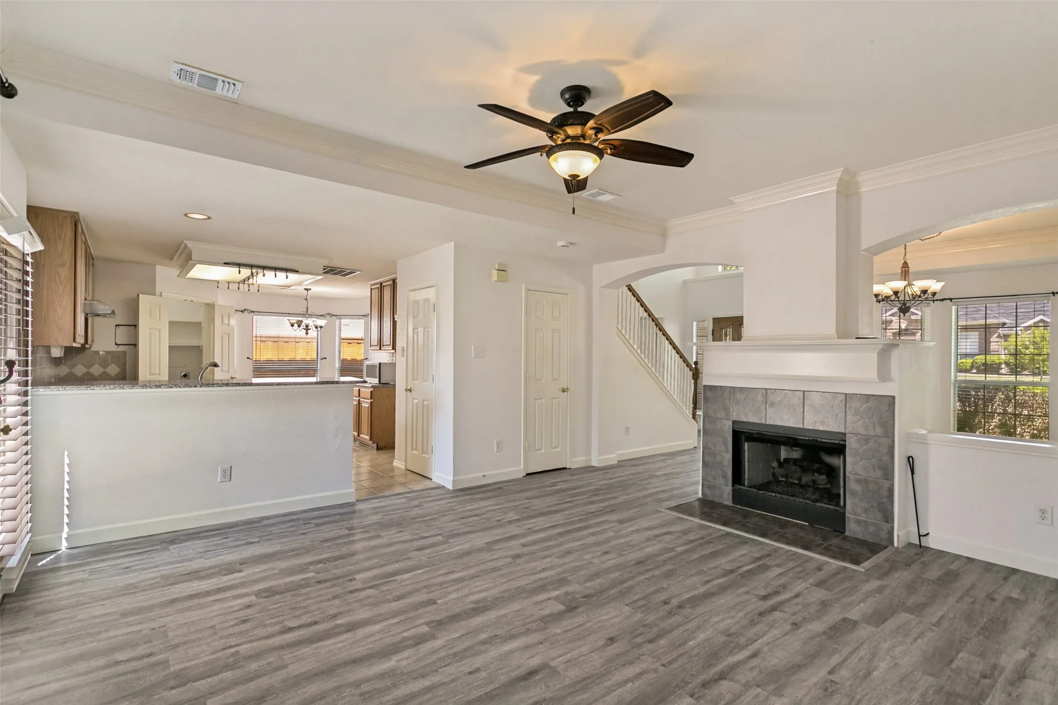 Wood-finish flooring throughout, featuring a tiled fireplace with mantel, a ceiling fan with integrated lighting, and an open concept to the kitchen area