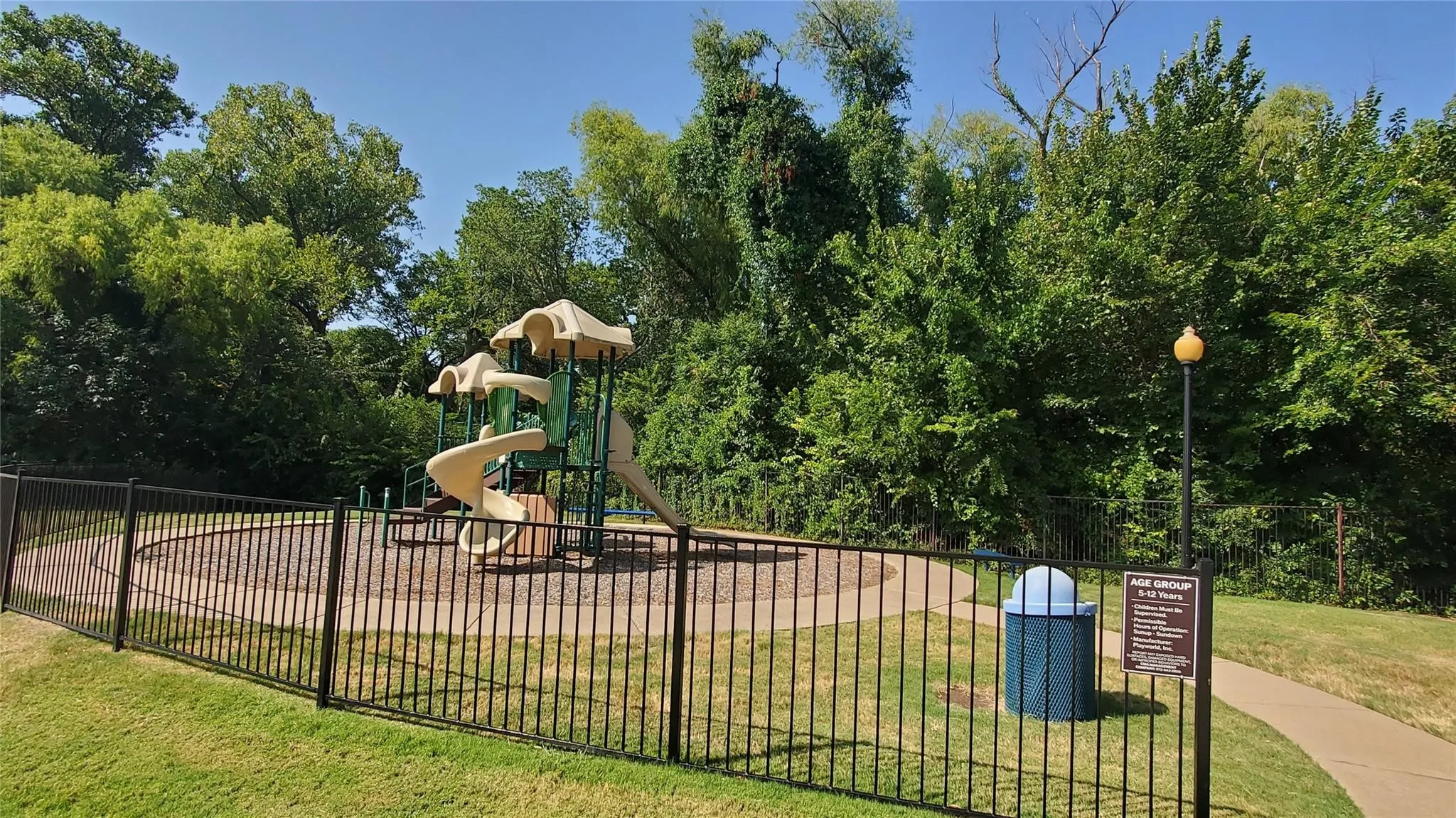 Fenced playground with multiple slides, surrounded by mature trees, featuring a paved walkway