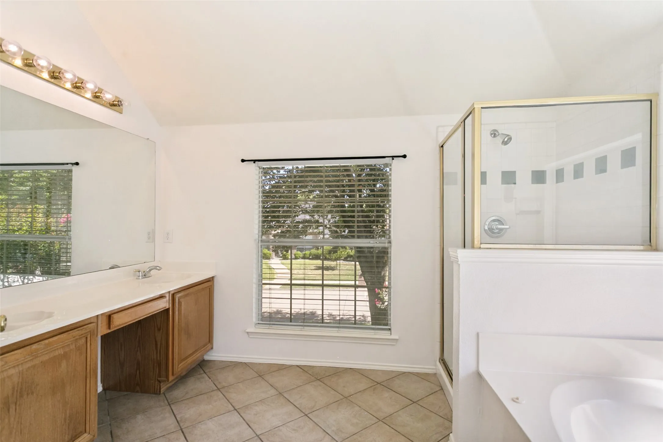 Vaulted ceiling with overhead lighting, dual vanity with wood-finish cabinetry, tile flooring, and a glass-enclosed shower
