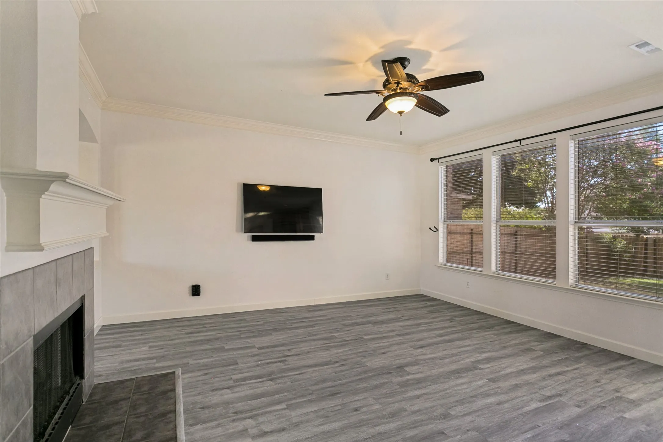 Spacious living area featuring plank-style flooring, a tile-surround fireplace with mantel, crown molding, a ceiling fan, and multiple windows