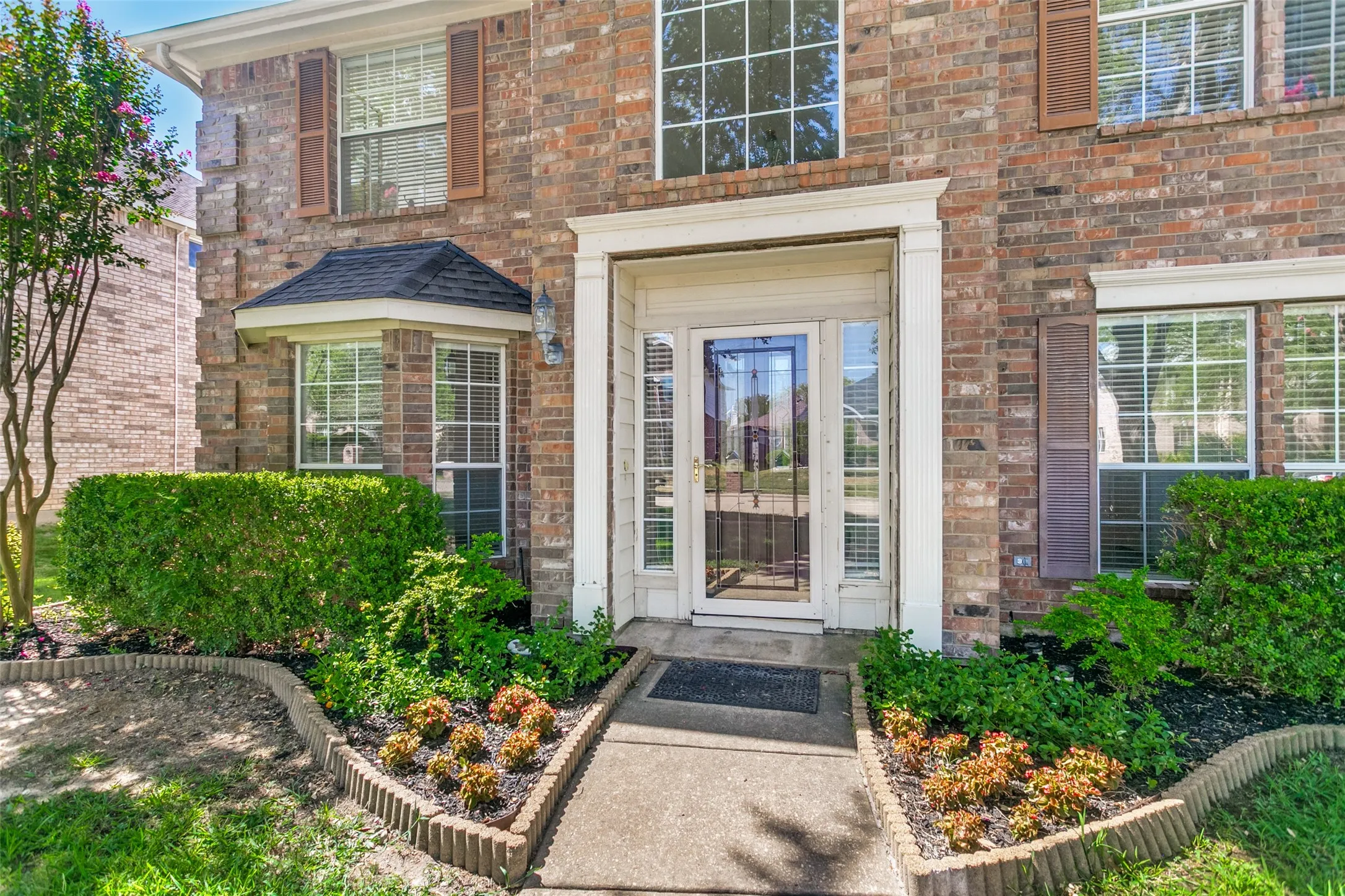 Brick facade with contrasting shutters, prominent bay window, landscaped front yard, and a white-framed entry door with a sidelight