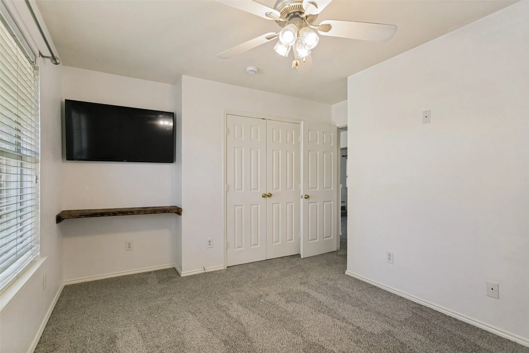 Neutral tone wall paint, textured carpet flooring, ceiling fan with light fixture, louvered closet doors, and window with blinds