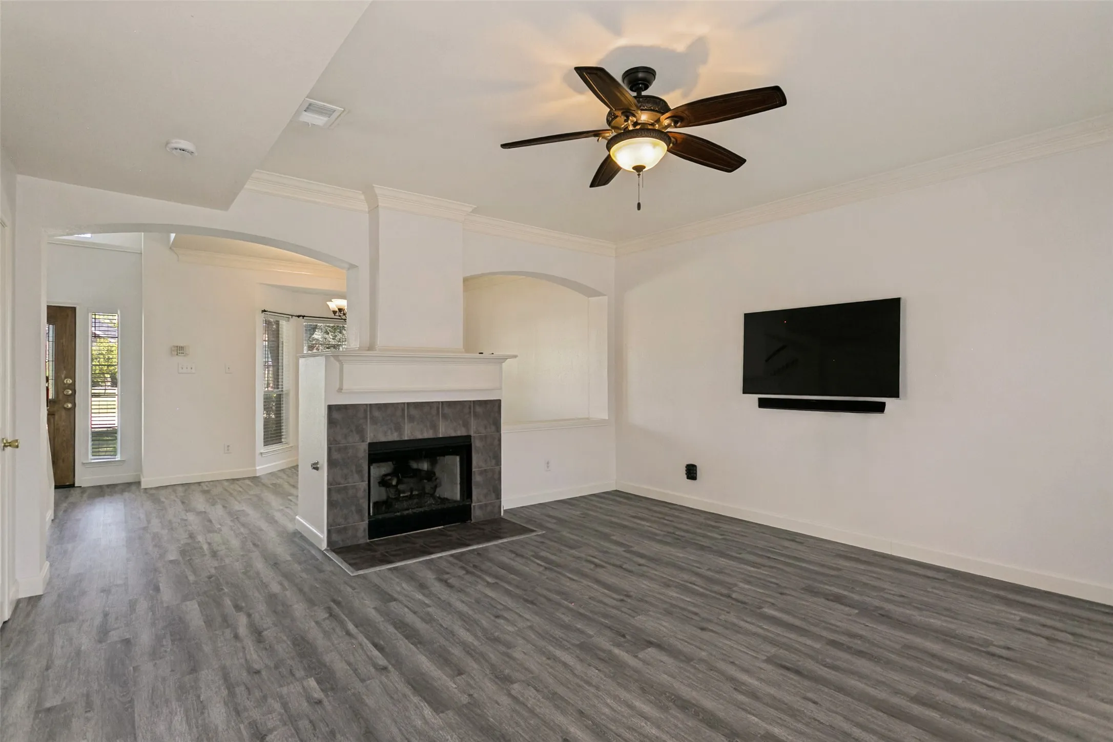 Living area featuring wood-finish flooring, a tiled fireplace, crown molding, arched doorways, and a ceiling fan with integrated lighting