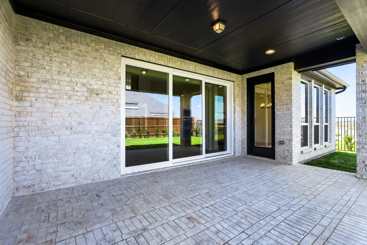 Covered outdoor patio with paver flooring, brick exterior, white-framed sliding glass doors, and a black-framed glass-paneled door